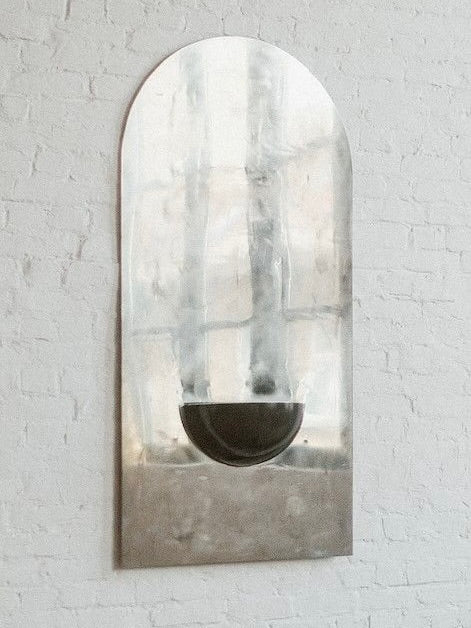 The Mirror of Simple Souls (Large) by COSEINCORSO is a contemporary steel mirror with a minimalist arched design, featuring abstract gray accents and a black semicircle, handmade in Belgium and shown on a white brick wall.
