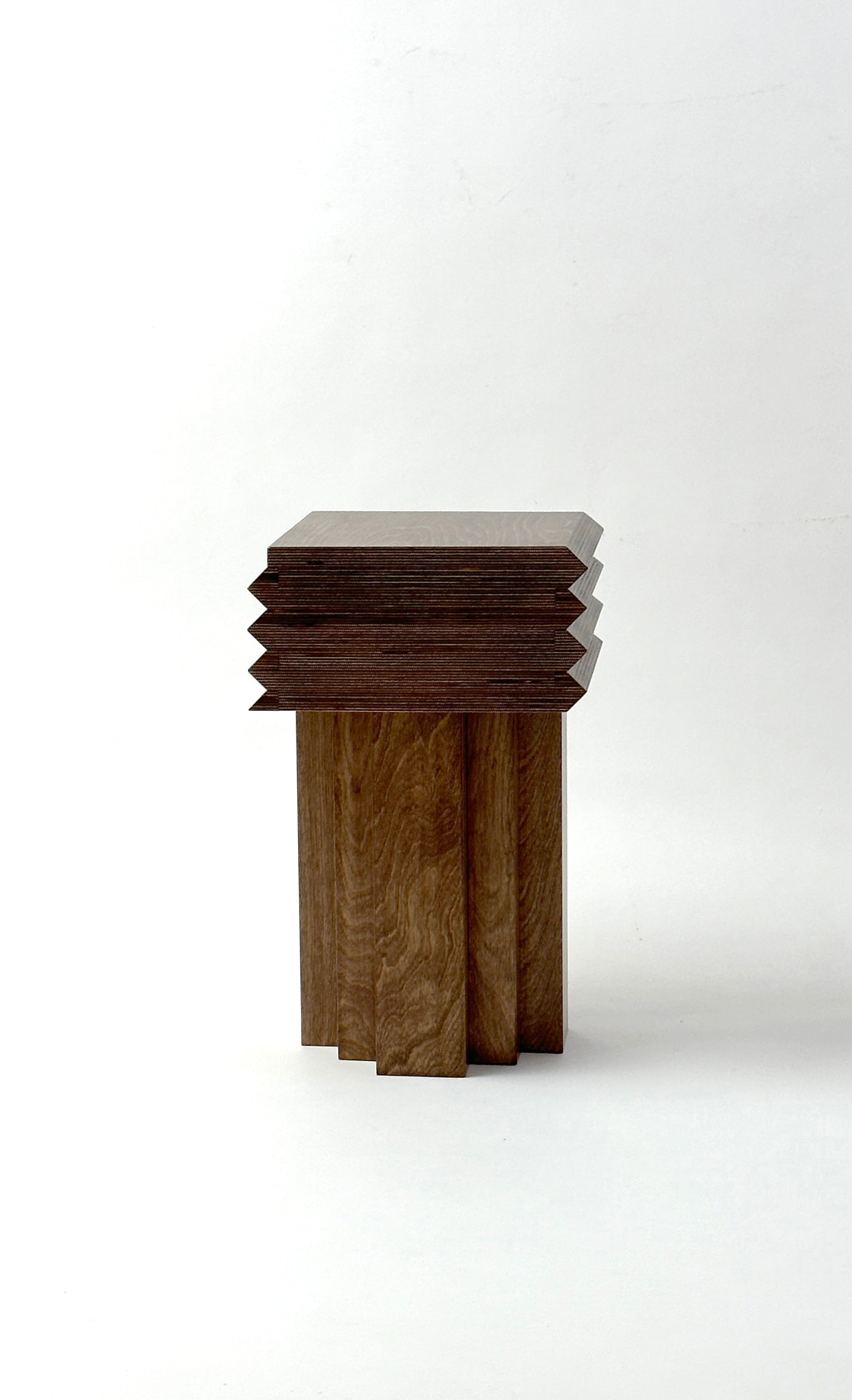 The MM Stool - Dark Oak by GOONS is a modern wooden side table with a square top, geometric base, and sharp edges, crafted by the Paris design studio GOONS. Finished in rich brown, it exemplifies artisanal furniture artistry.