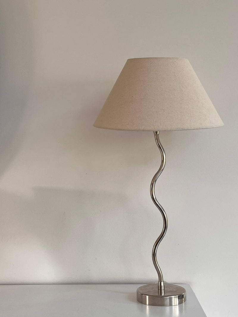 The Maud Lamp - Nickel by Maud Vaughan features a beige fabric shade and wavy metallic base, standing on a white surface against a light wall.