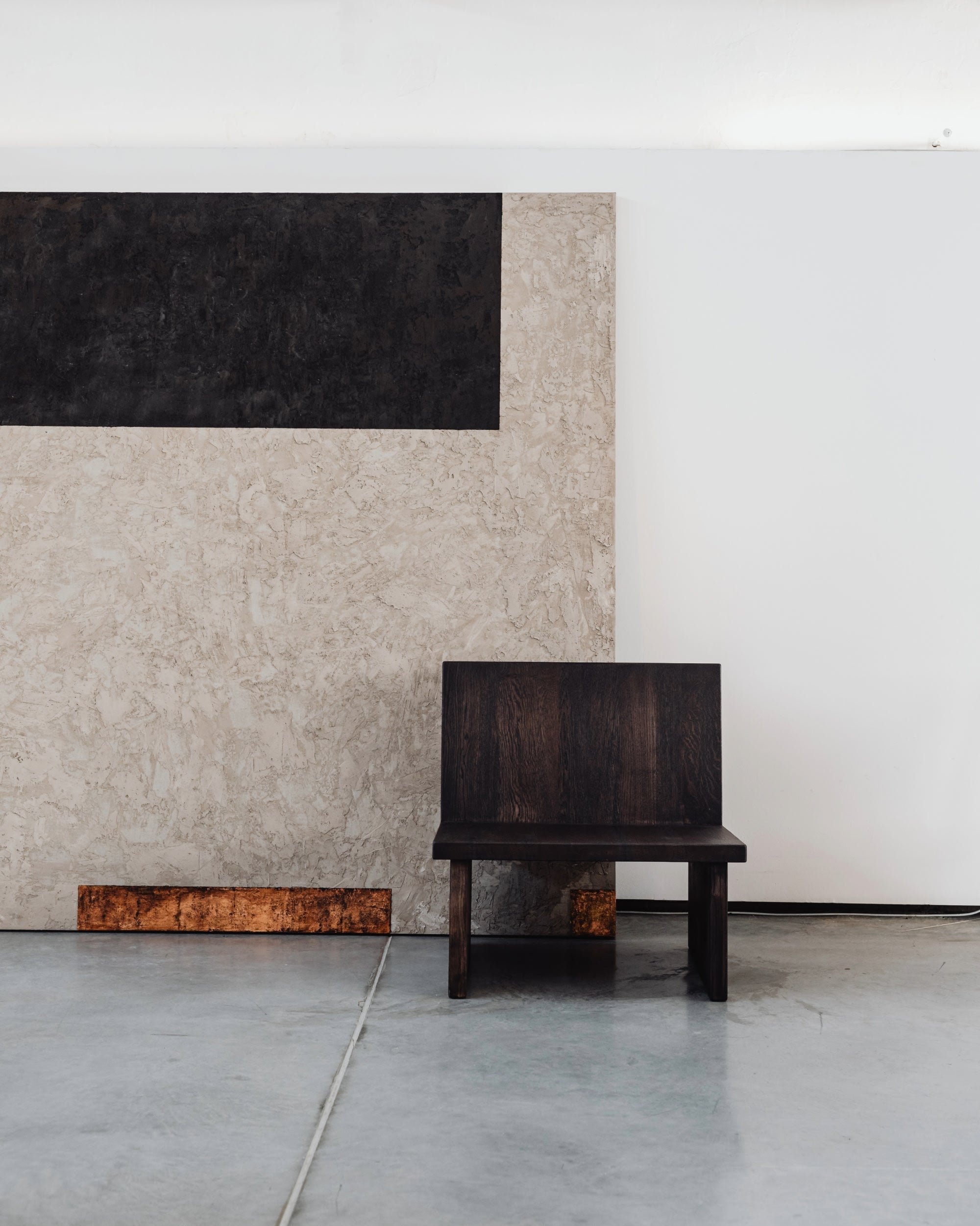 A minimalist scene features the ZEBU Chair by un'common, crafted from solid oak with a natural oil wax finish, placed on a concrete floor beside a large beige and black textured panel against a plain white wall.