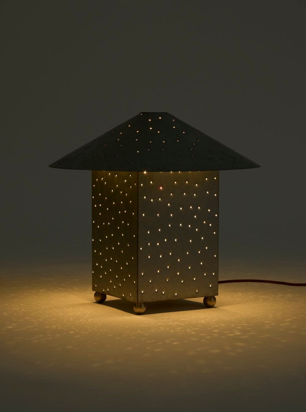 The Lin Table Lamp, Steel from the MUHLY brand boasts a modern hand-punched metal construction with a rectangular base and a broad, conical shade adorned with countless small holes. This design creates a captivating starry-night effect on the surface below. The lamp emits a warm, soft glow and has an exposed cord for plugging in.