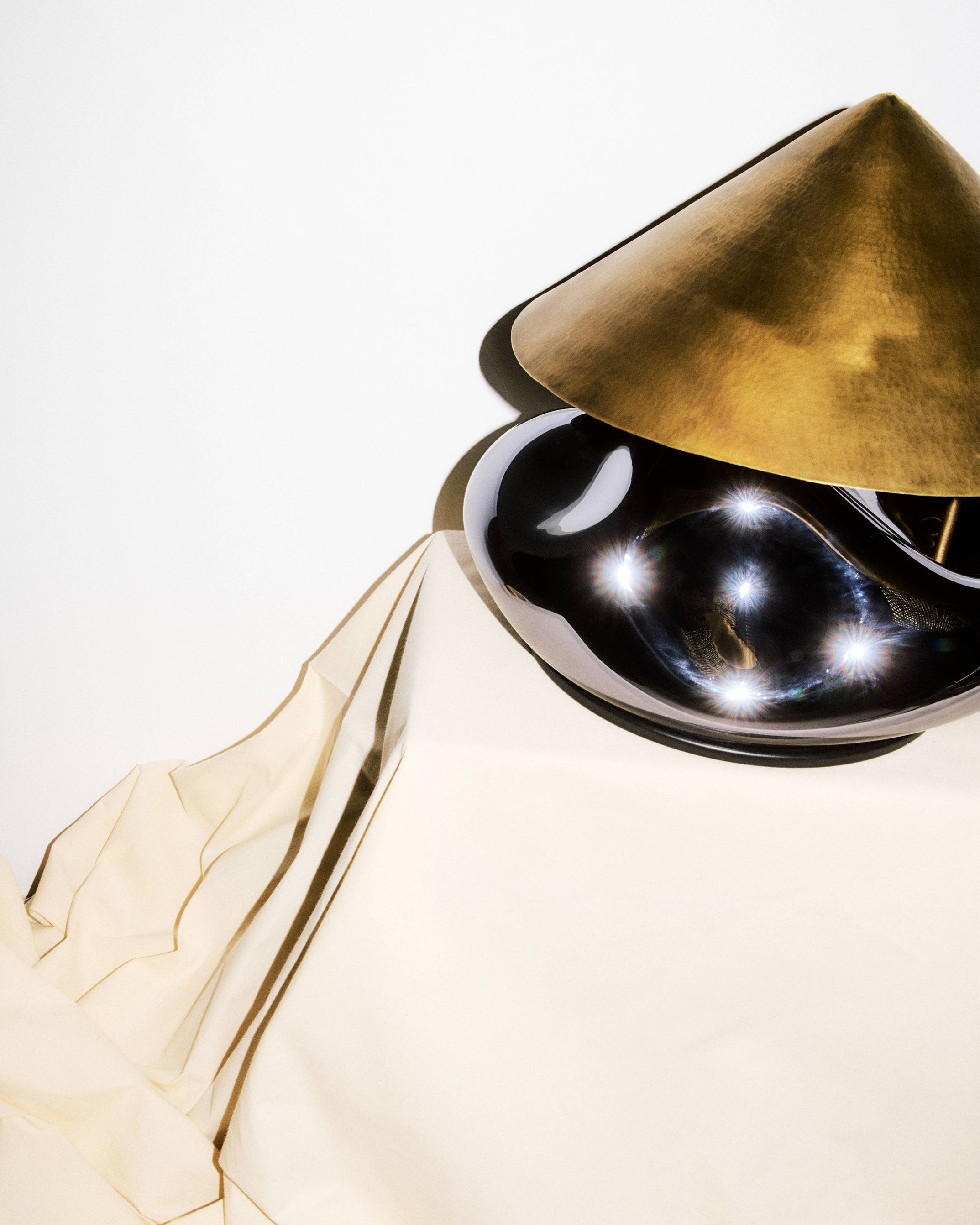 The HOMA Obsidian Lamp features a shiny, dark base and a metallic gold cone-shaped shade, set on cream-colored fabric against a white background, creating elegant reflections and highlights.