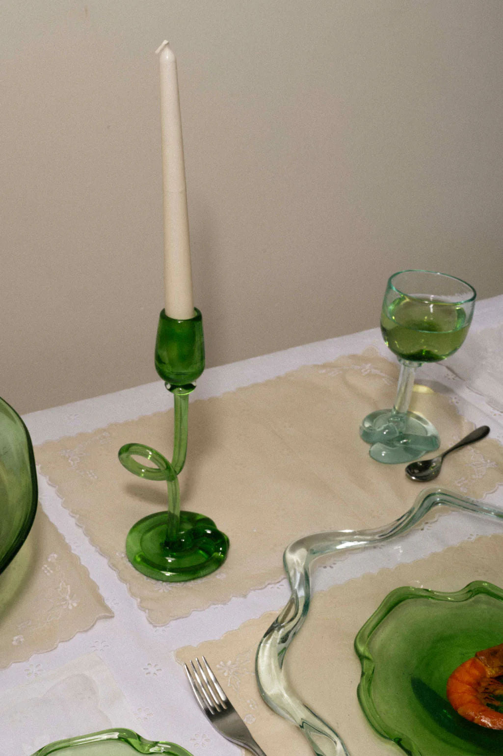 A table setting with the Thaw - Recycled Glass Candleholder in Clear by Completedworks holding a white candle, green glass goblet, silverware, and uniquely shaped green glass plates on a white tablecloth with beige placemats.