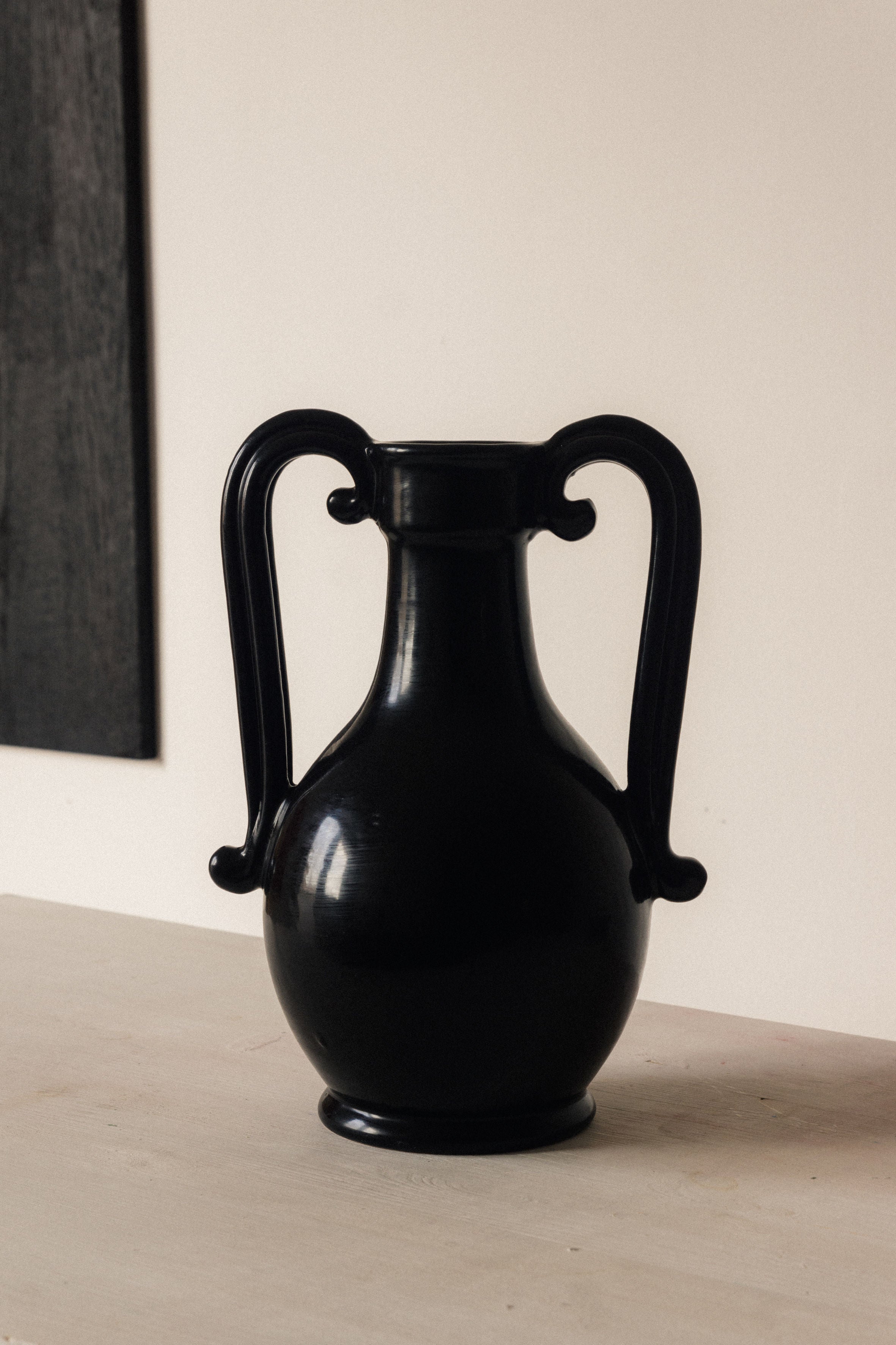 The Fiasca - Minimalist Vase in Ceramic by COSEINCORSO, handmade in Italy, features a glossy black finish, two curved handles, and a rounded body with a narrow neck. It sits on a light surface under soft lighting against a neutral background.