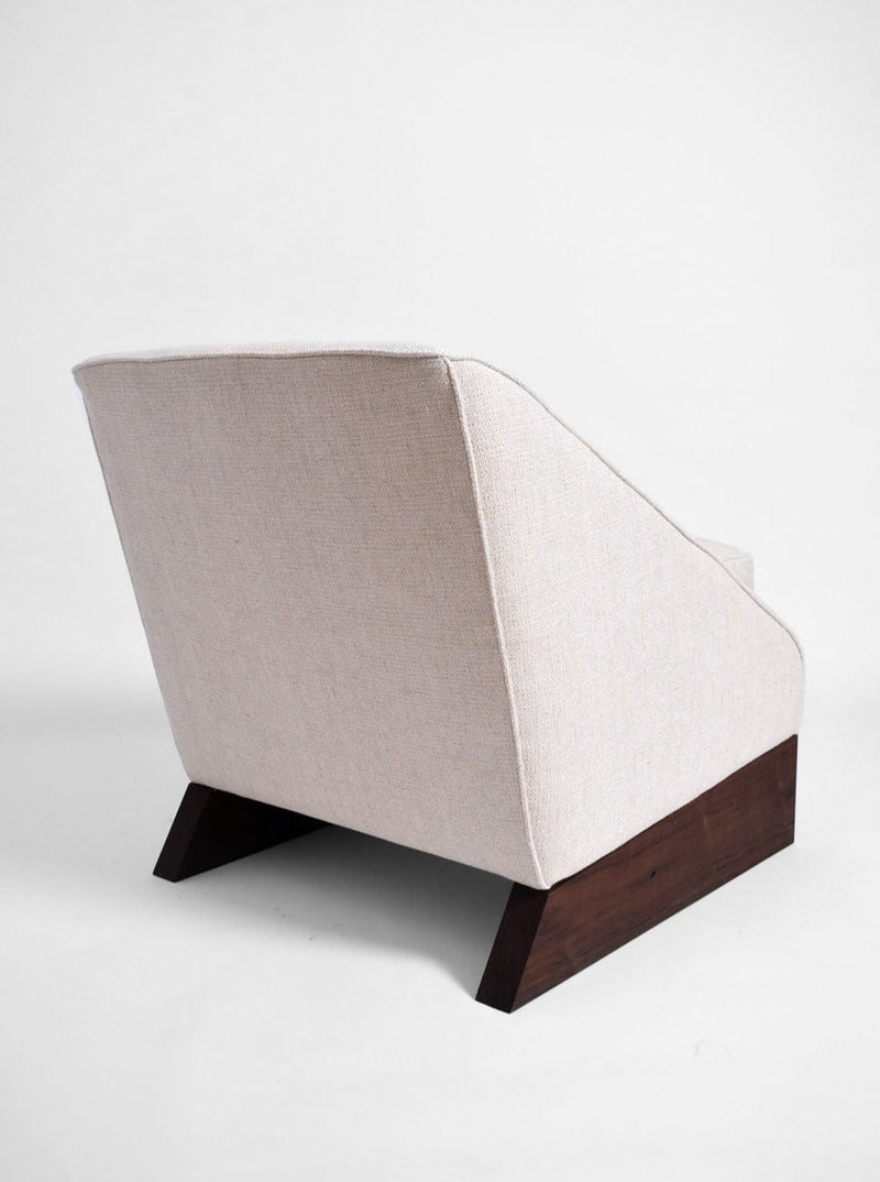 The Arrábida Armchair by Barracuda Interiors features light beige upholstery and angled dark wooden legs, highlighting its sleek modern design from the back against a plain white background.