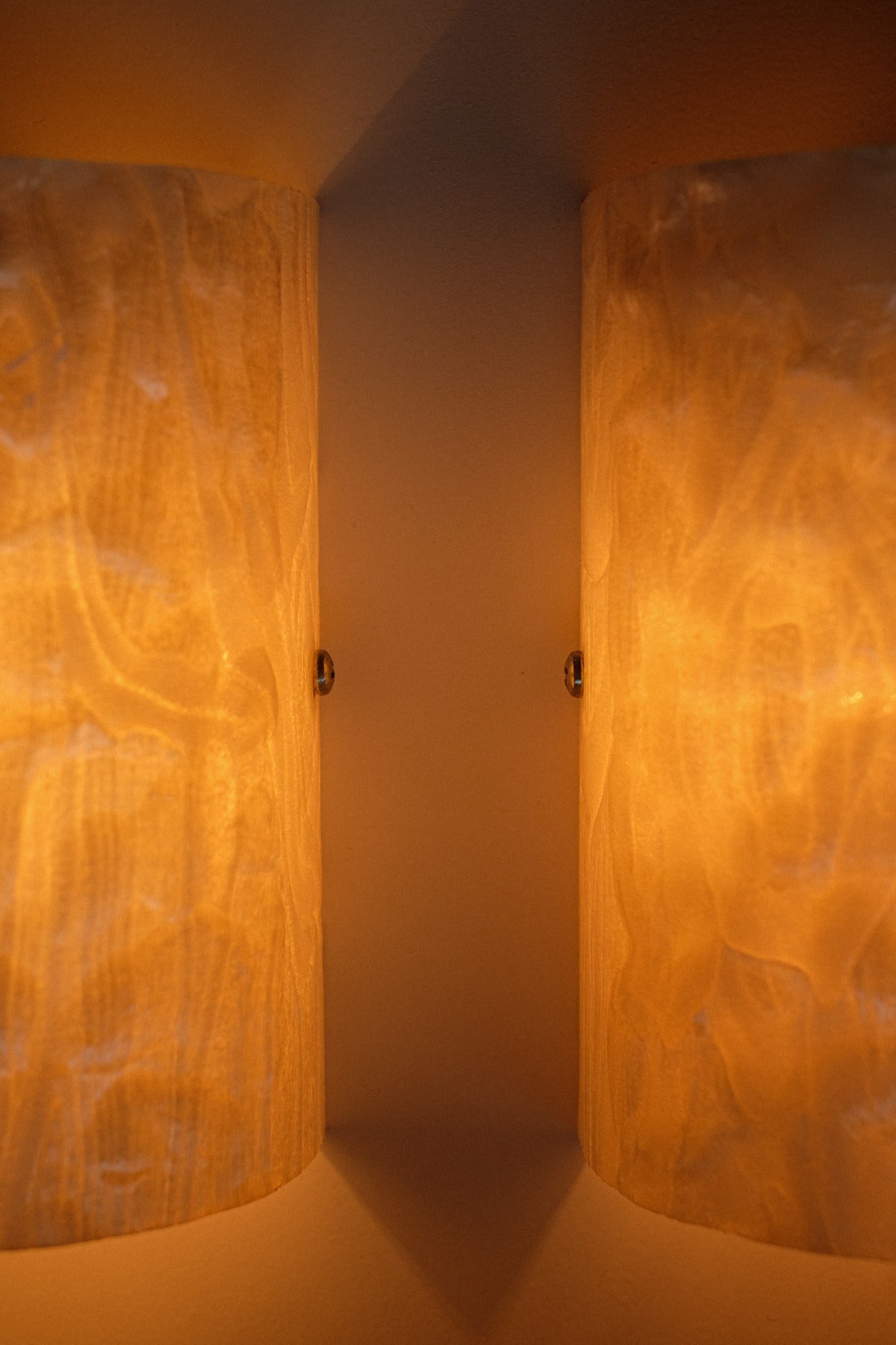 The Set of 2 Mother-of-pearl Wall Lamps by septembre studios features cylindrical designs with textured surfaces, emitting a warm yellow glow on either side of a corner for a cozy, inviting ambience.