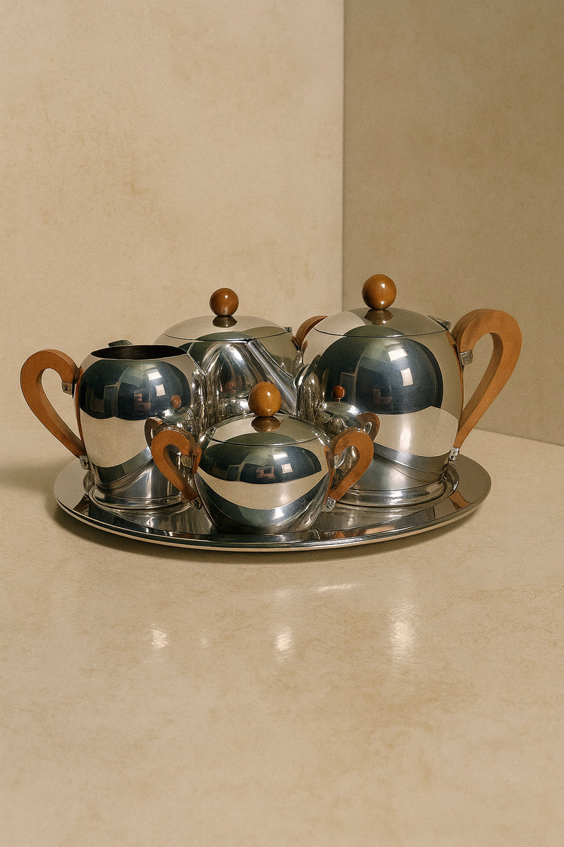 The Art Deco Tea/Coffee Set ‘Bombè’ by Alessi from Dodo Vintage, featuring reflective silver pieces with wooden handles, is displayed on a round tray against a beige background, highlighting its subtle Art Deco design.