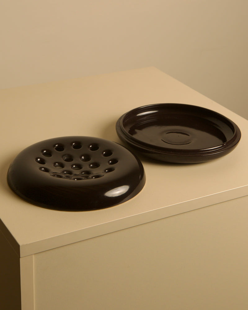 Two round, dark brown glazed ceramics from Treaptyque sit side by side on a beige surface—one is an Italian Brown Ceramic Ashtray by Angelo Mangiarotti for Brambilla (1960s) with multiple holes; the other is a shallow dish with a central circular indentation.