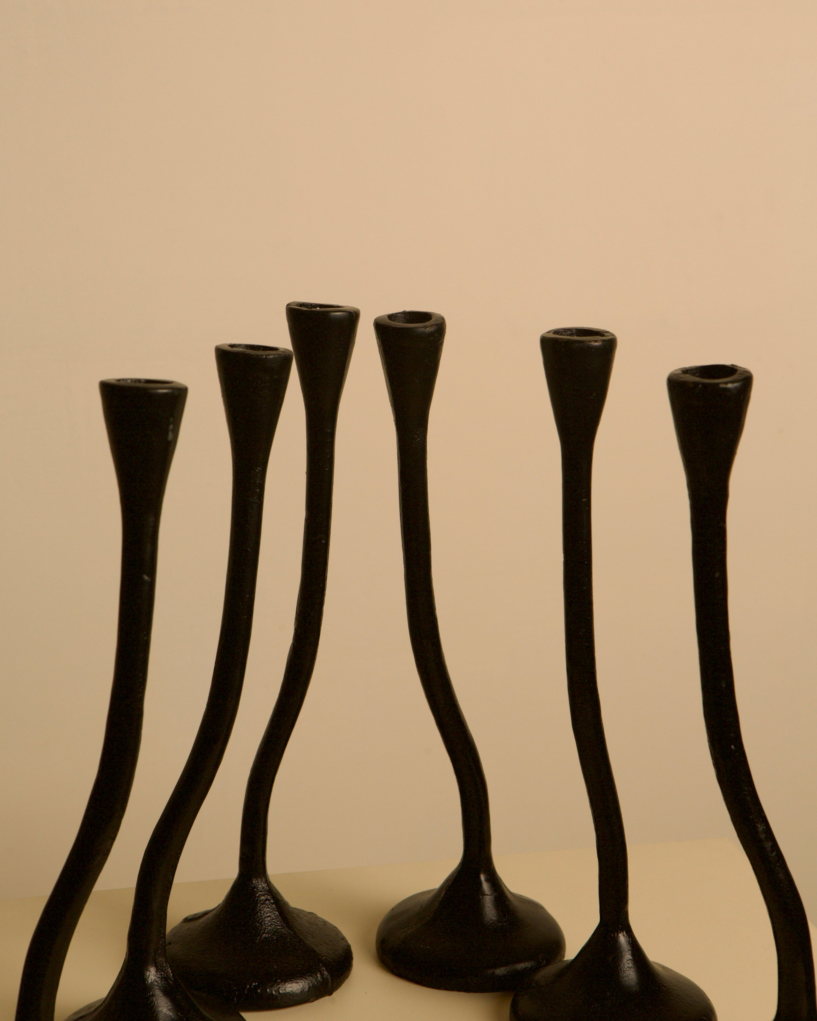 A set of 5 "Twisted" black cast aluminum candleholders by Treaptyque, featuring slender, curved 1980s-inspired designs with flared bases and narrow tops, stand on a light surface against a beige background.