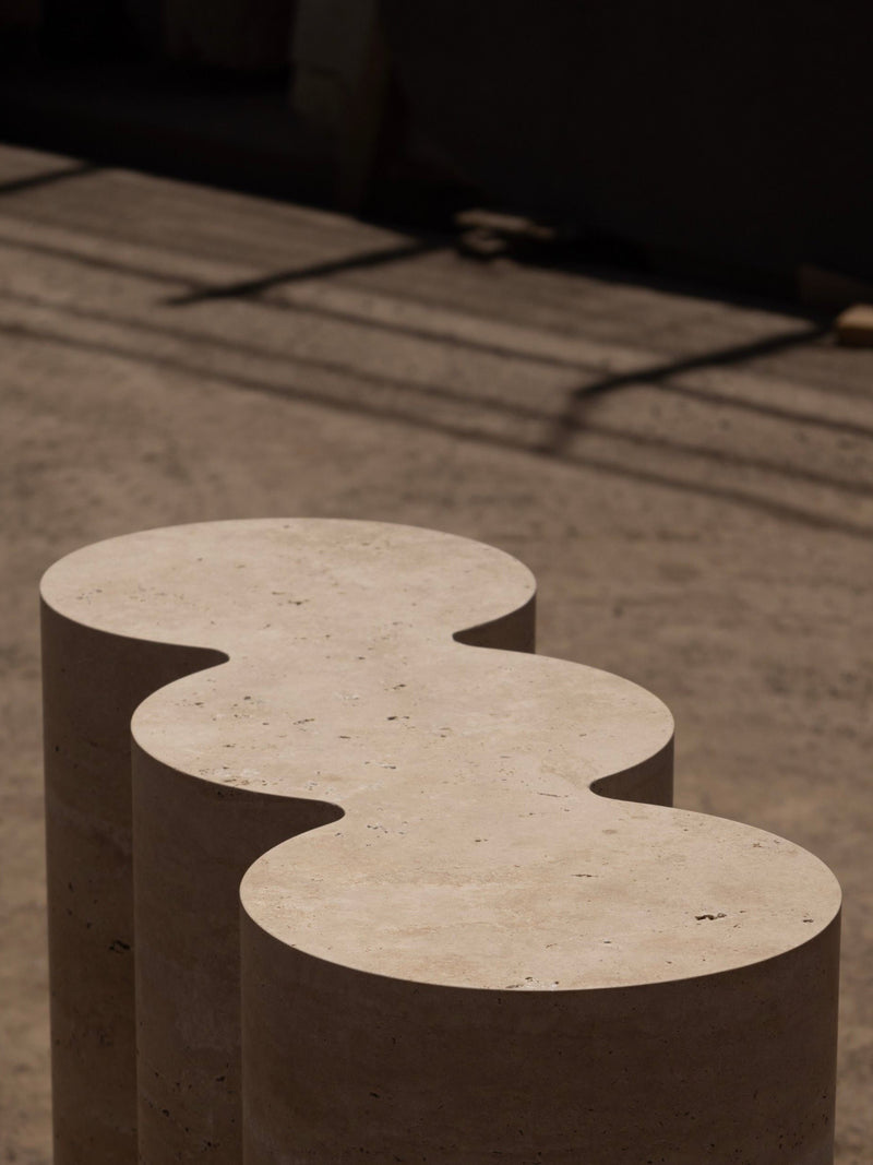 Three beige, cylindrical stone columns with scalloped edges define Monolith’s M_008 Travertine Console, their triple-column geometry casting soft shadows across sunlit sand.