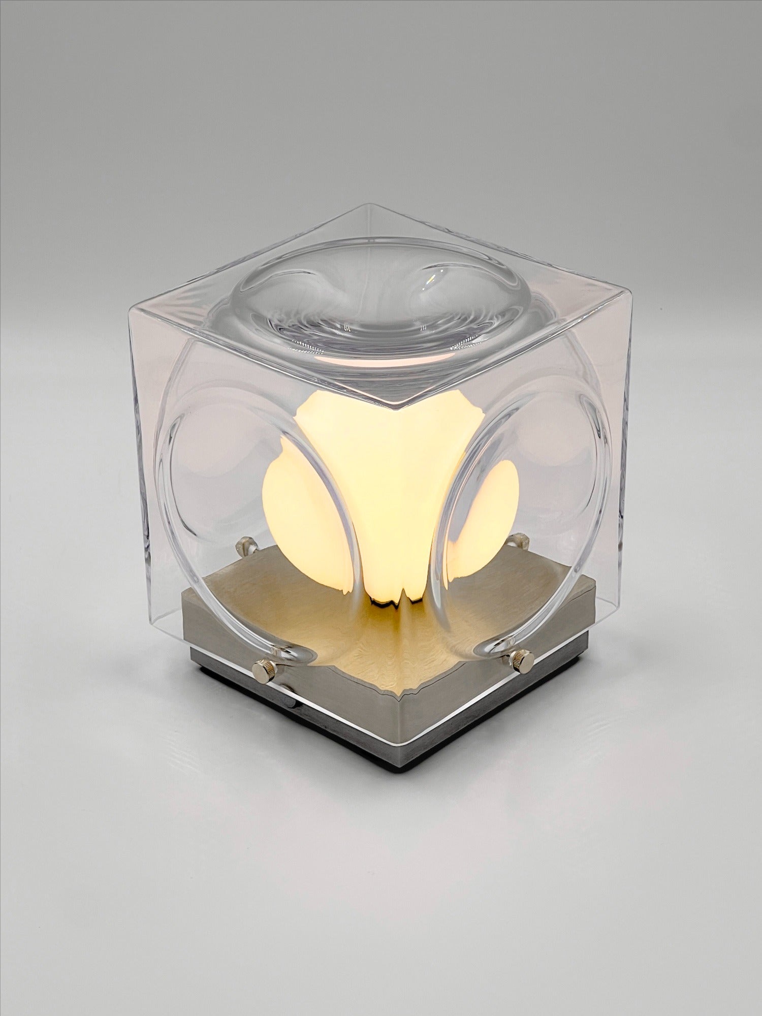 The Chant Portable Table Lamp by Lee Broom features a hand-blown glass cube with a transparent, glossy finish and rounded indents, diffusing warm light from its center, set atop a metallic square base.