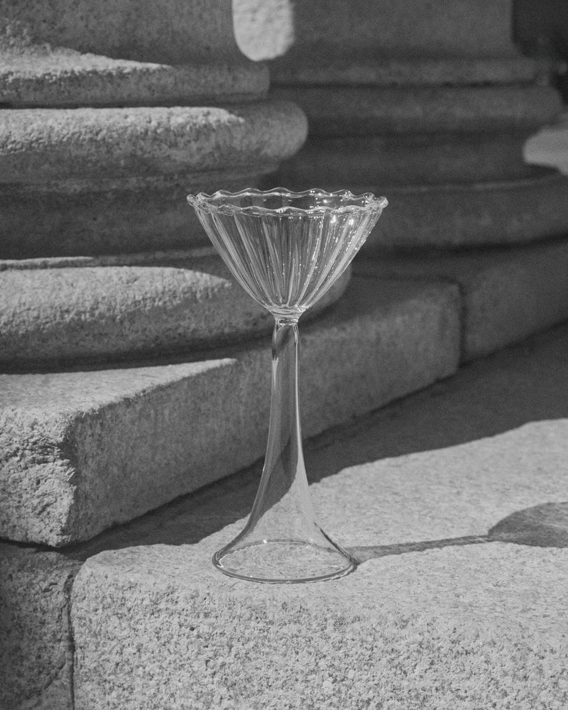 The Campanula — Clear Cocktail Glass by Agustina Bottoni stands on stone steps in sunlight, set against textured columns. A black-and-white photo highlights the elegant handmade borosilicate glass artistry from Italy.