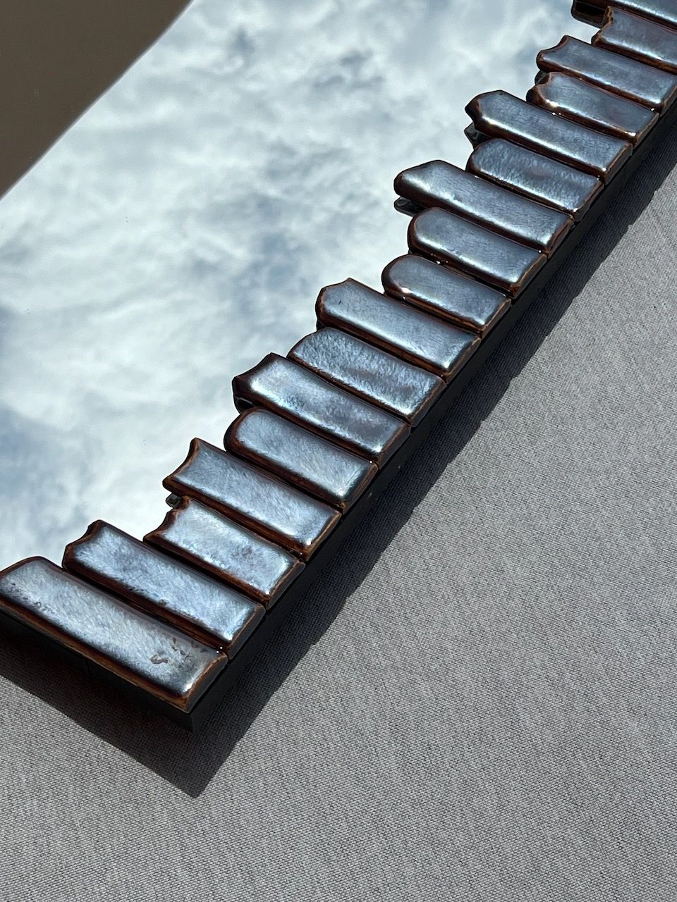 A mirror from the Katia Langeard Cliff Collection features a wavy ceramic edge in metallic blue, resting on gray fabric and reflecting a cloudy sky—seamlessly merging nature with contemporary design.