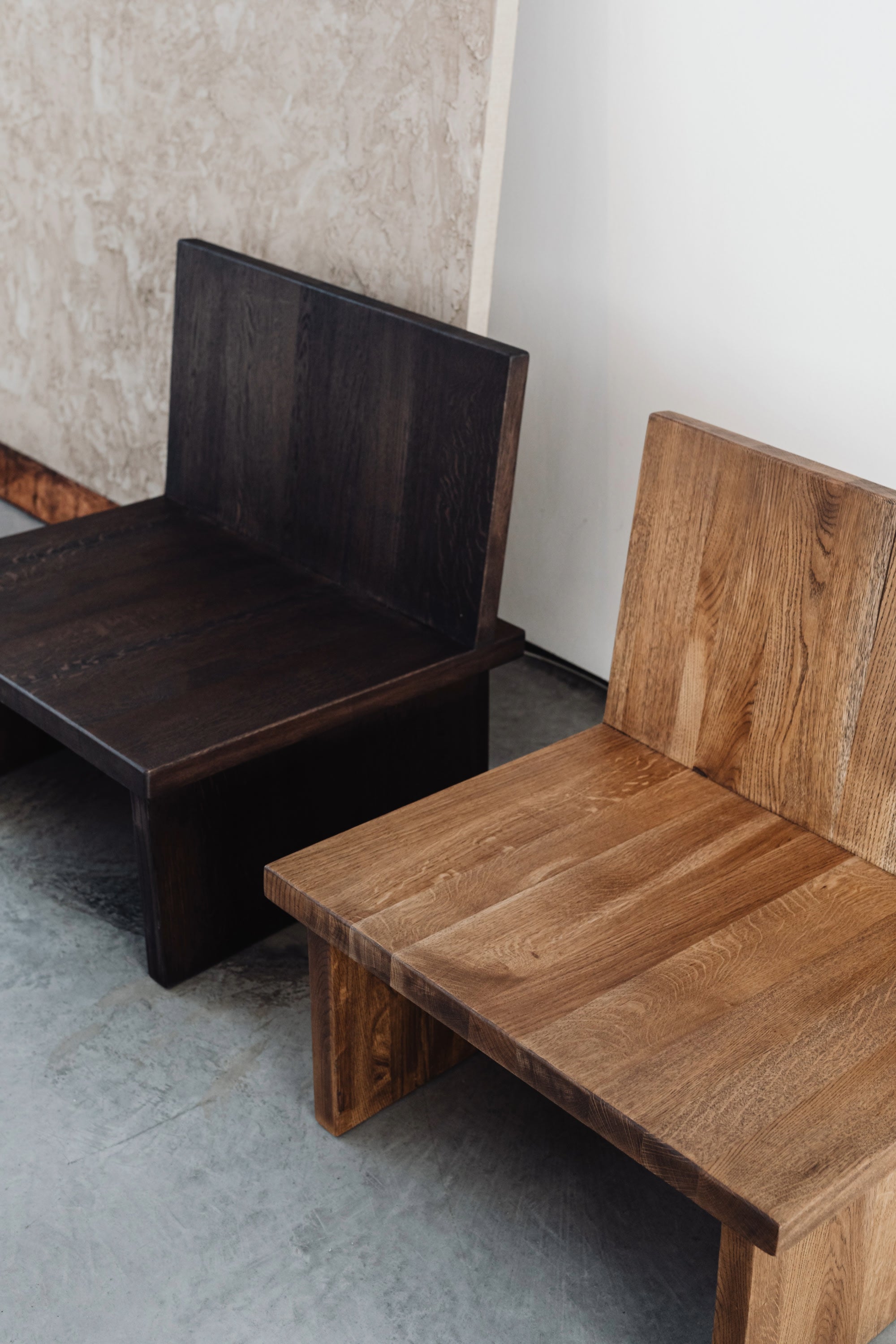 Two ZEBU Chairs by un'common—solid oak with natural oil wax finish, one dark brown and one light brown—stand side by side on a concrete floor, set against textured and white walls in the background.