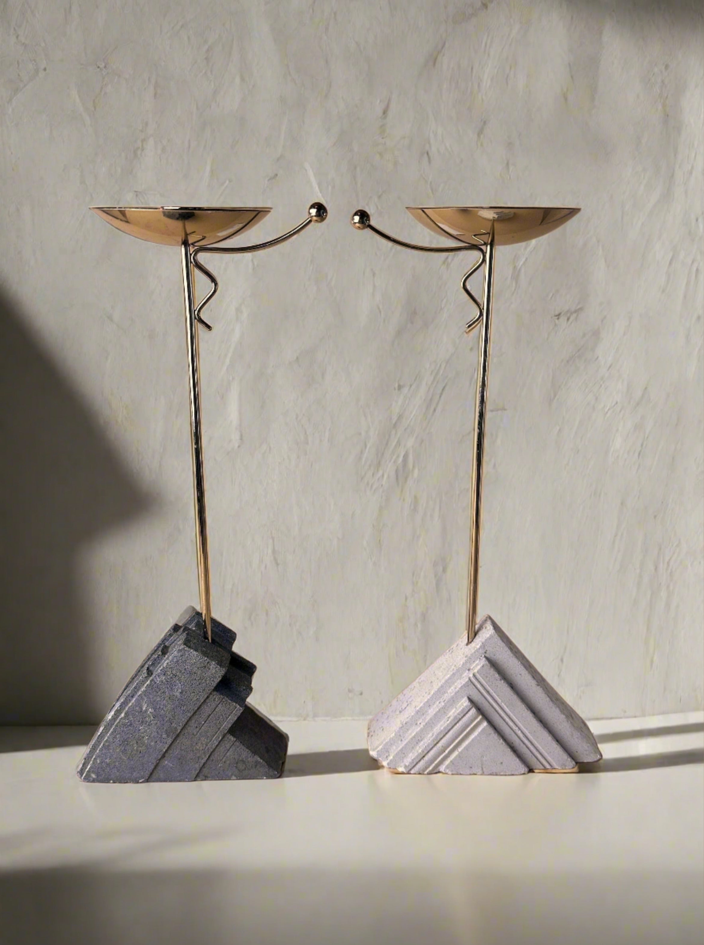 The Art Deco Revival candlesticks by Dodo Vintage feature geometric stone bases and slender brass stems with metal dishes, displayed side by side against a neutral textured wall.