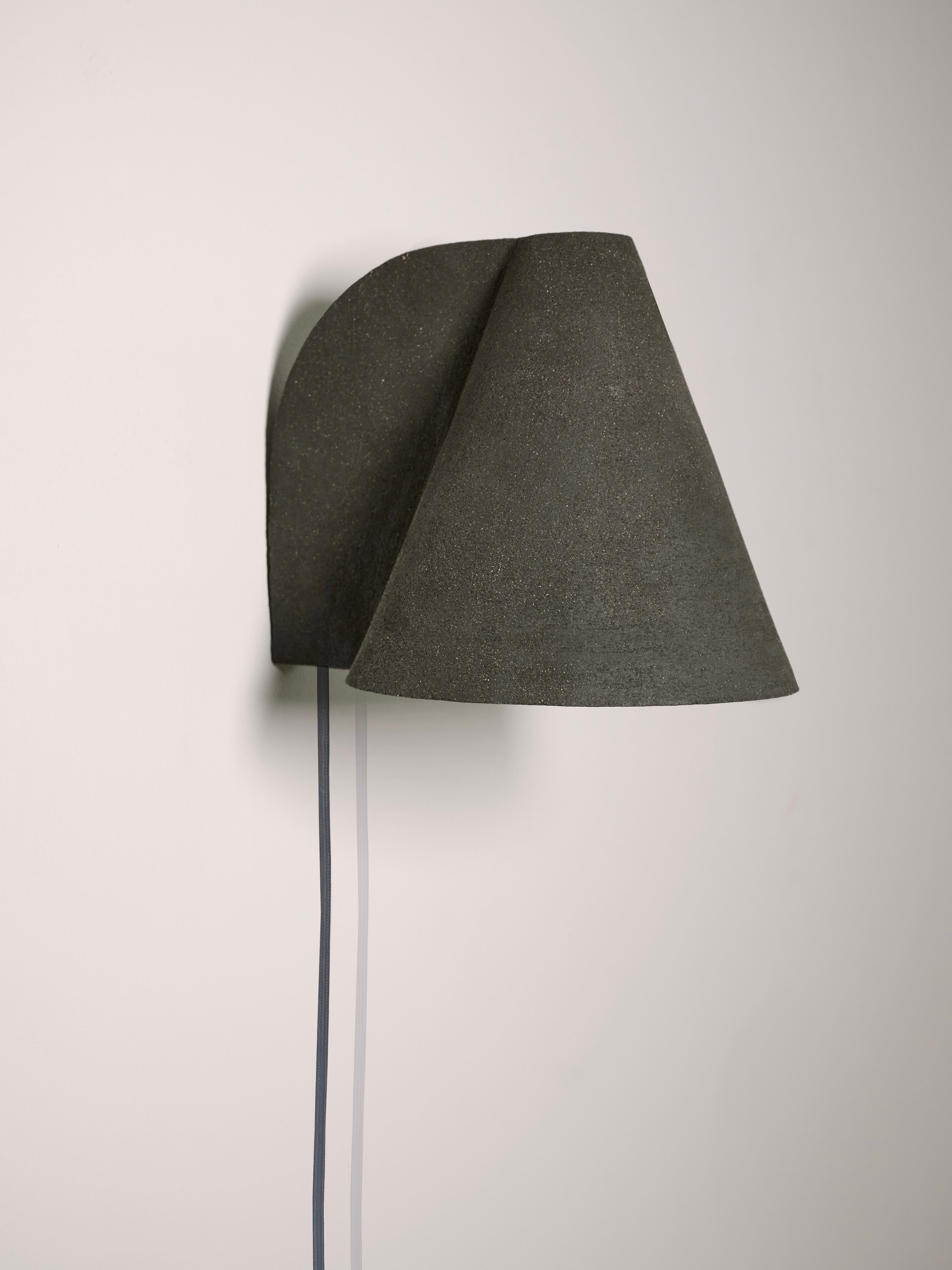 The Esoteric Wall Lamp by Mariza Galani features a minimalist black design with a conical shade and visible cord, mounted on a white wall for a simple yet striking look.