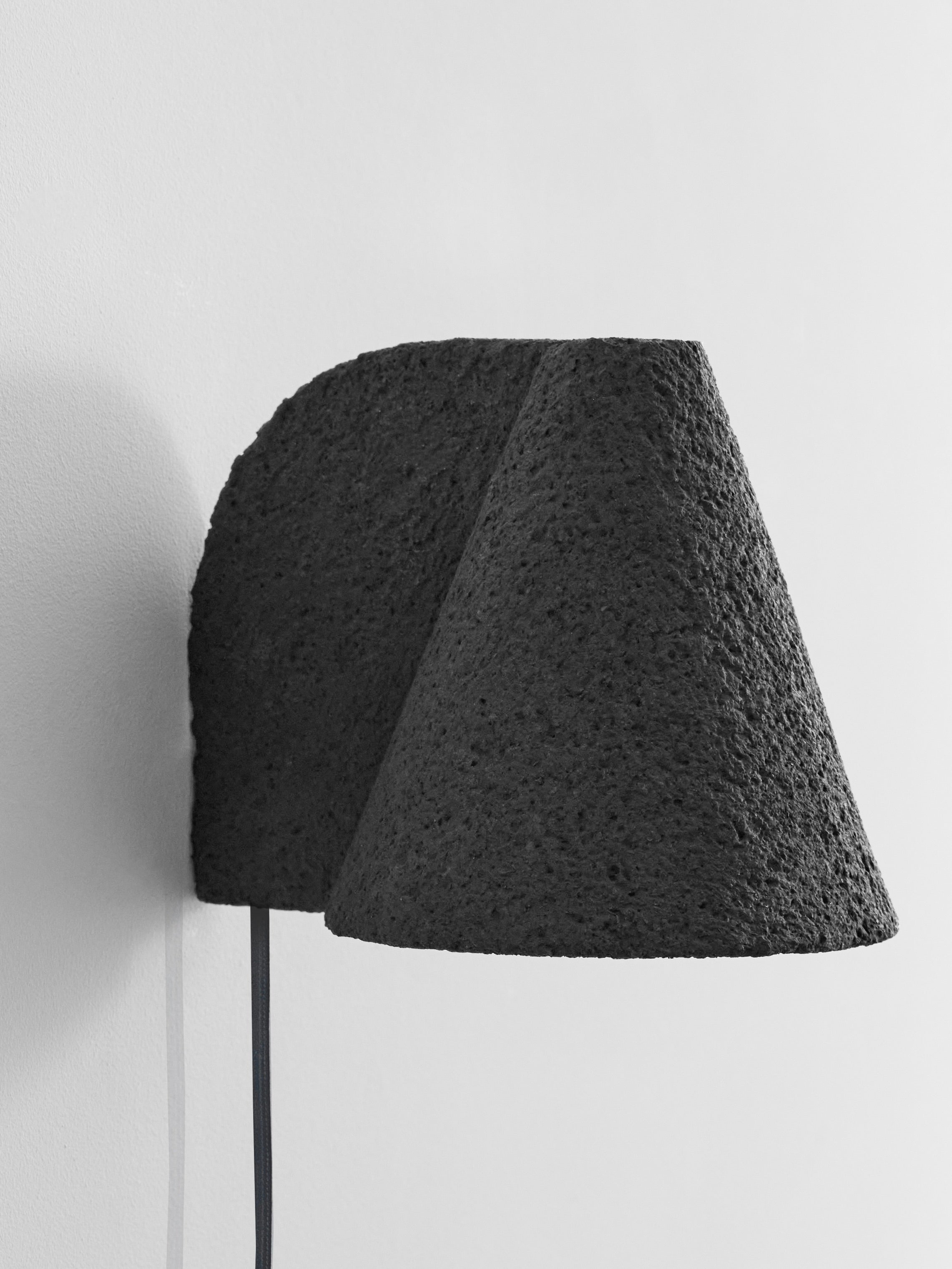 The Esoteric Granular Wall Lamp by Mariza Galani features a textured black volcanic rock shade and matching curved base, mounted on a white wall with a distinctive black cord for unique, modern lighting.