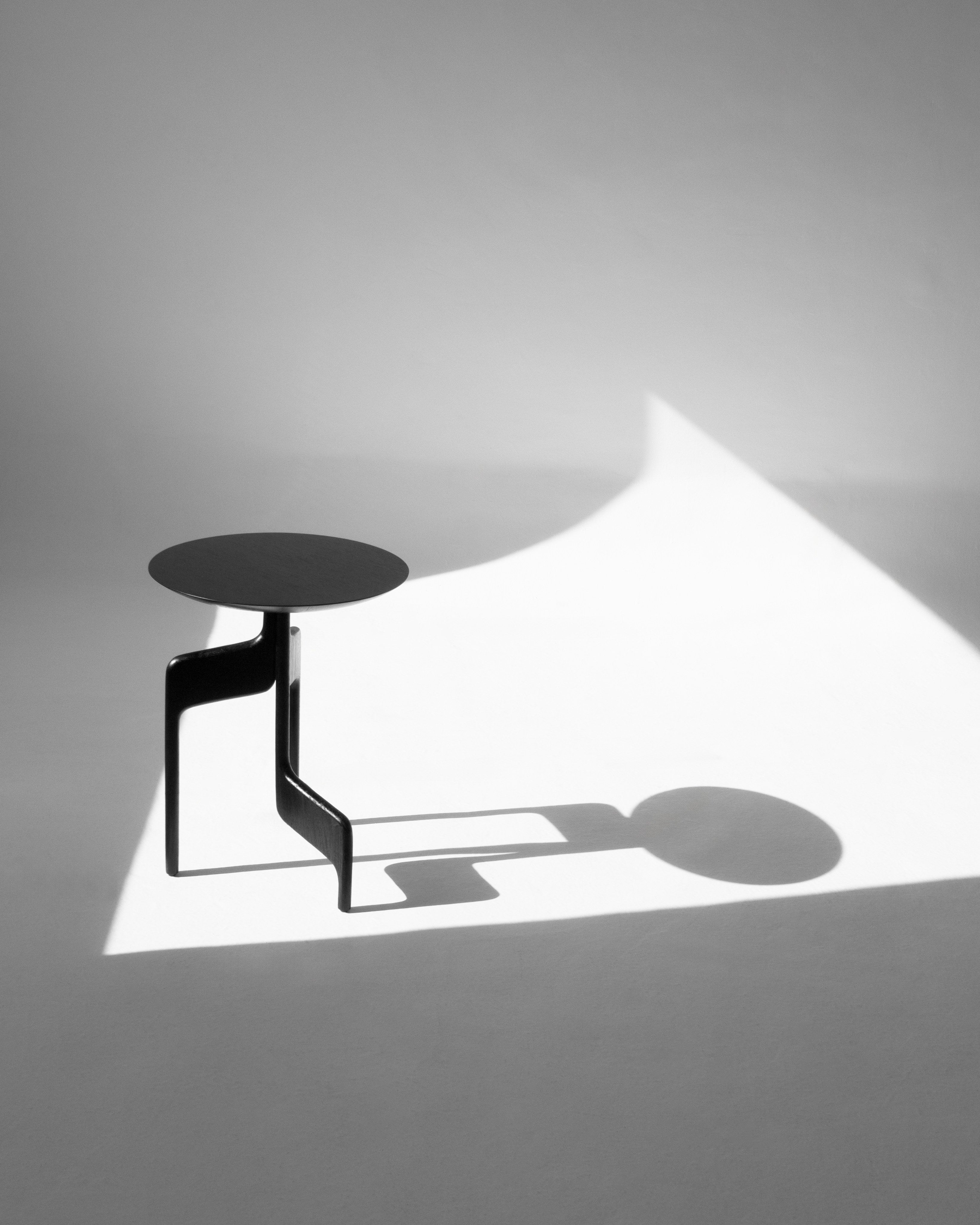 The "Vereda" Side Table by Luciano Costa Estúdio, in a modern minimalist black design, stands on a white floor with a bold shadow, capturing the artistic elegance and high-contrast style of transformative accessories.