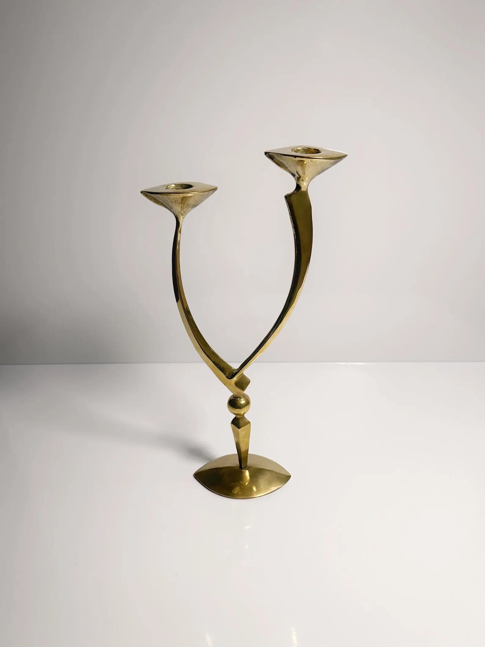 A Mid-Century Candleholder crafted by Dodo Vintage features two upward-curving arms in a polished brass finish, elegantly displayed on a simple white surface against a neutral backdrop. Its sleek and minimalist design exudes a Schiaparelli-inspired style, offering it a contemporary and artistic look.