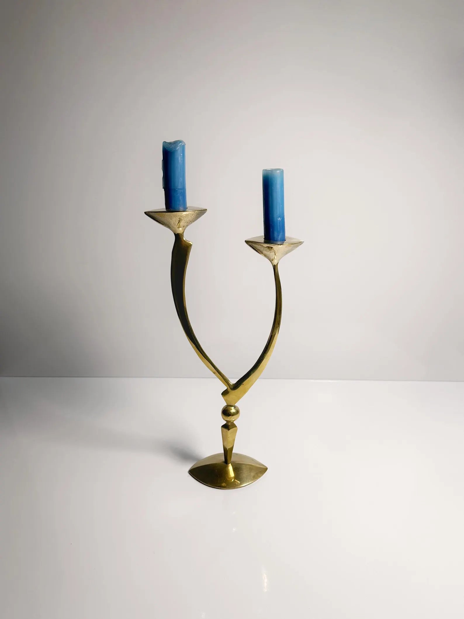 A Dodo Vintage Mid-Century Candleholder, featuring two arms each adorned with a blue candle, stands elegantly against a plain gray background. Its sleek, curved design in polished brass offers a symmetrical and sophisticated look. Beneath it lies a glossy white surface.