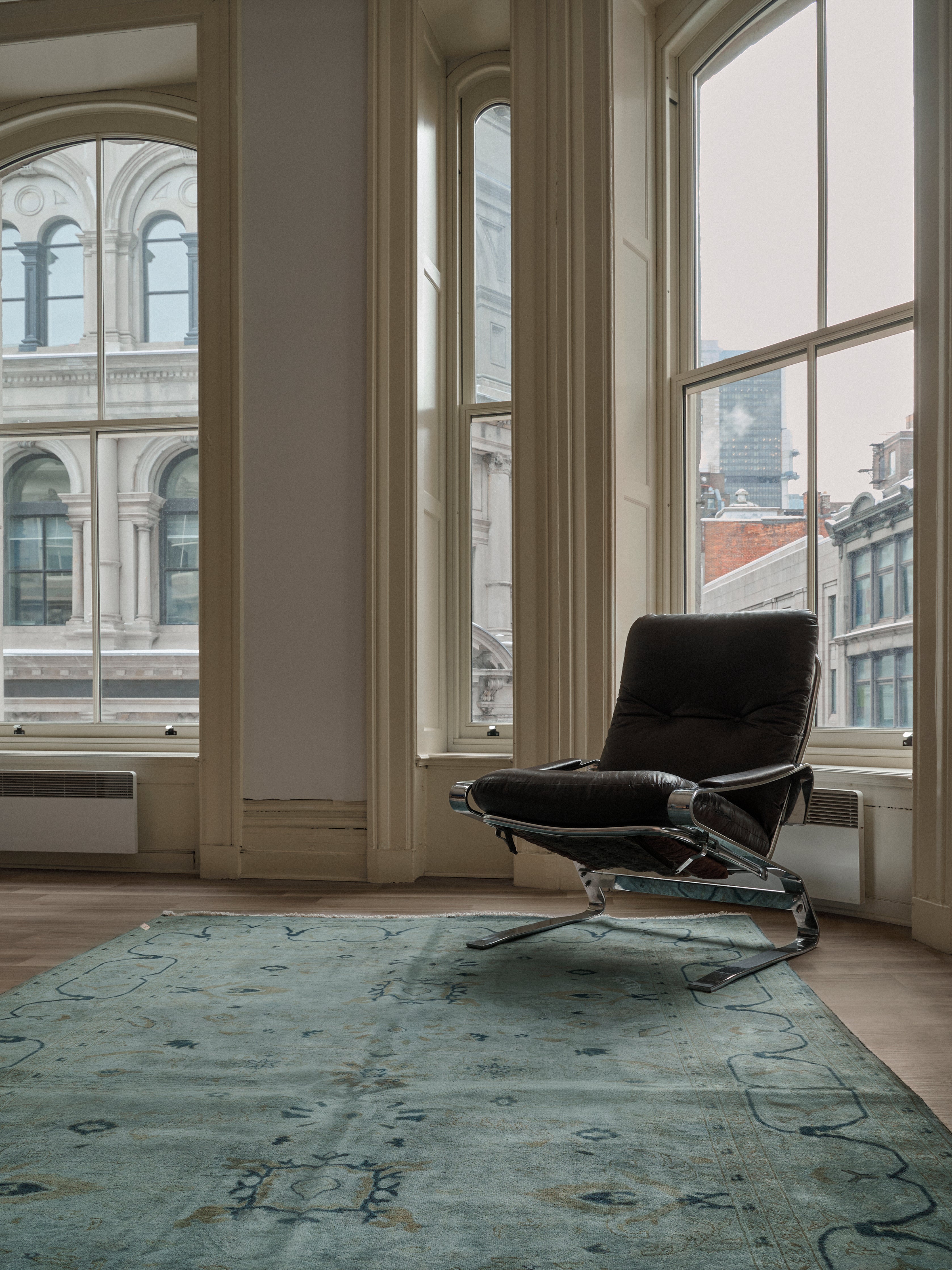 A modern interior showcases a sleek, dark chair with chrome legs atop an Overdyed Rug in Indigo by catalog:three. Large windows reveal classic architectural buildings against the city skyline, while natural light emphasizes the room's elegant design.