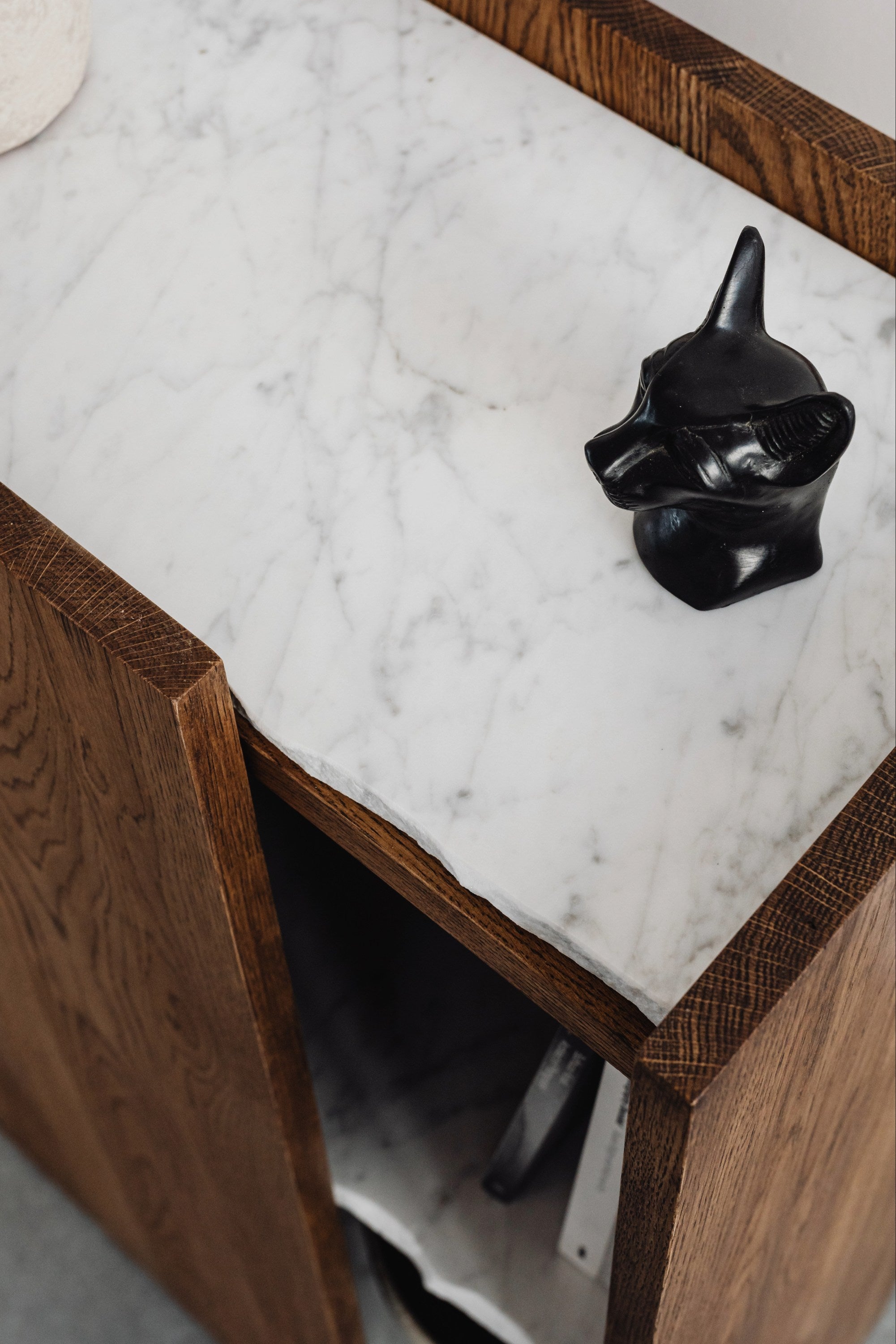 The PAVO Cabinet by un'common features solid wood construction with a white marble top, partially open to display a small black decorative cat figurine on its surface.