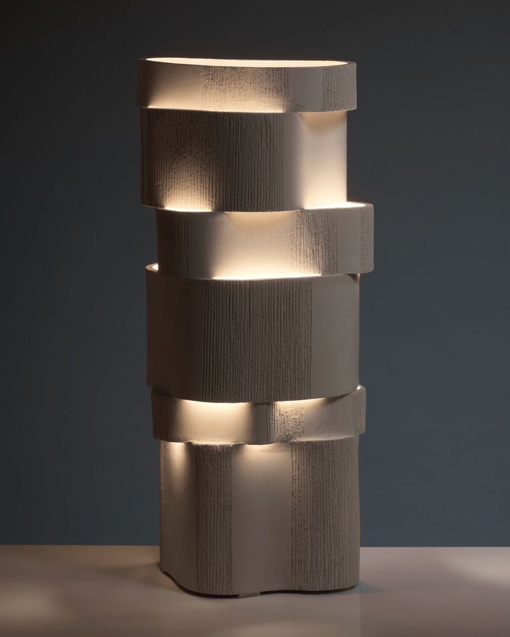 The 'Too 60' Ceramic Table Lamp by Katia Langeard features a modern, compact design with staggered layers that emit gentle light. Its textured matte beige finish adds warmth, perfect for display on any smooth surface against a dark backdrop.