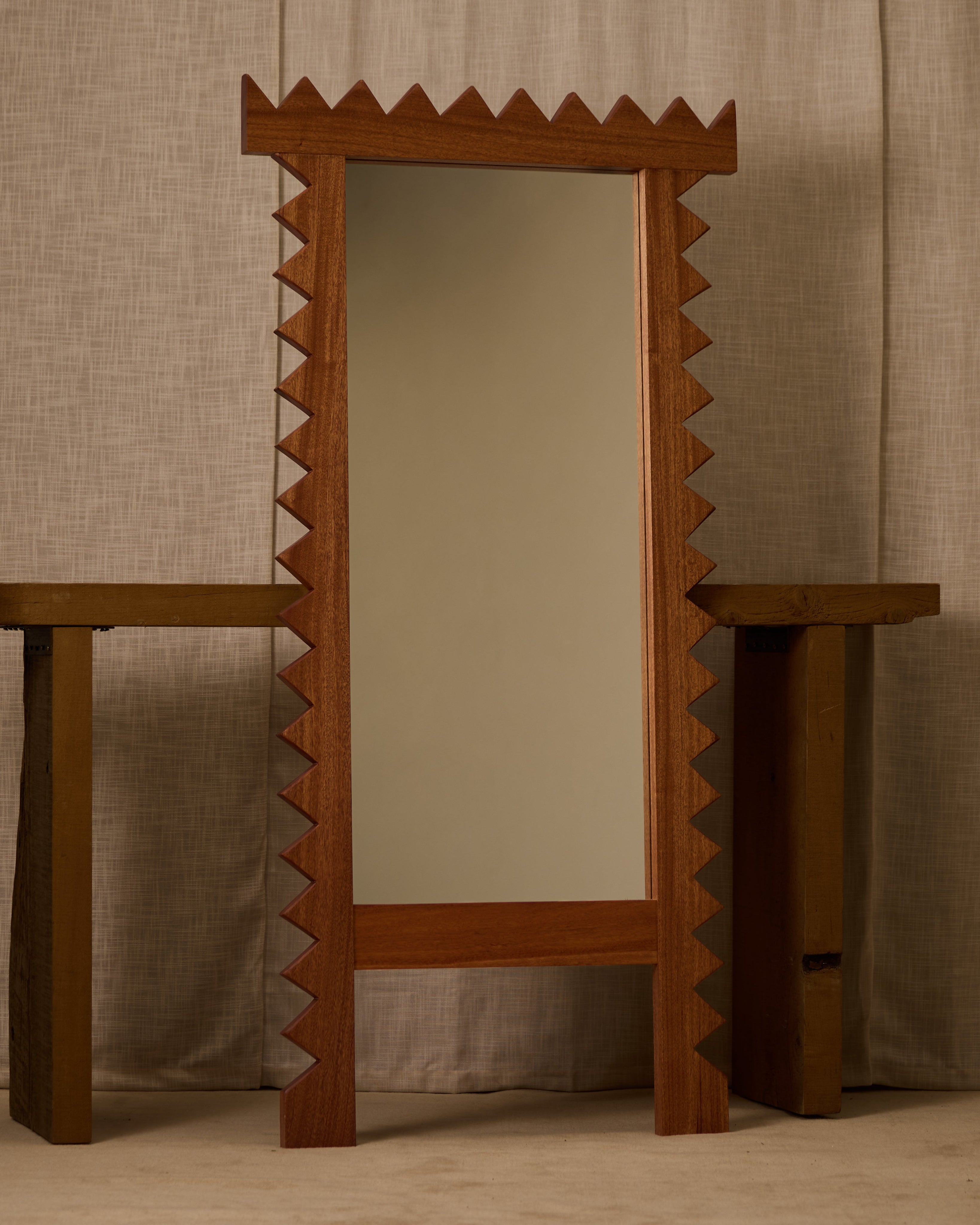 The Jura Mirror by Ah Um Design Studio, with its wooden frame and zigzag edges, stands between two rustic benches against a neutral backdrop, introducing bold geometric shapes to the space.