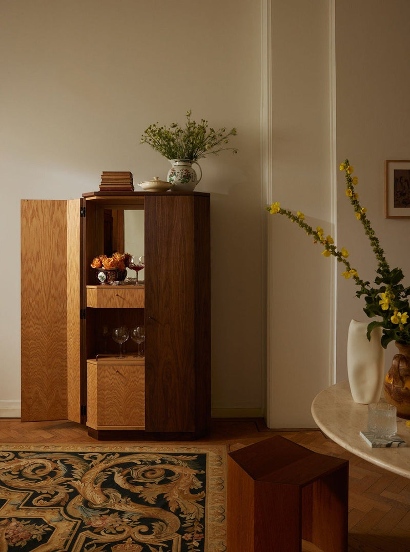 The Winston Drinks Cabinet in Walnut and Oak by Lemon stands open to reveal glassware and a vase. On top, a vase with greenery and stacked books sit, while nearby a white table with yellow flowers rests on an ornate rug.