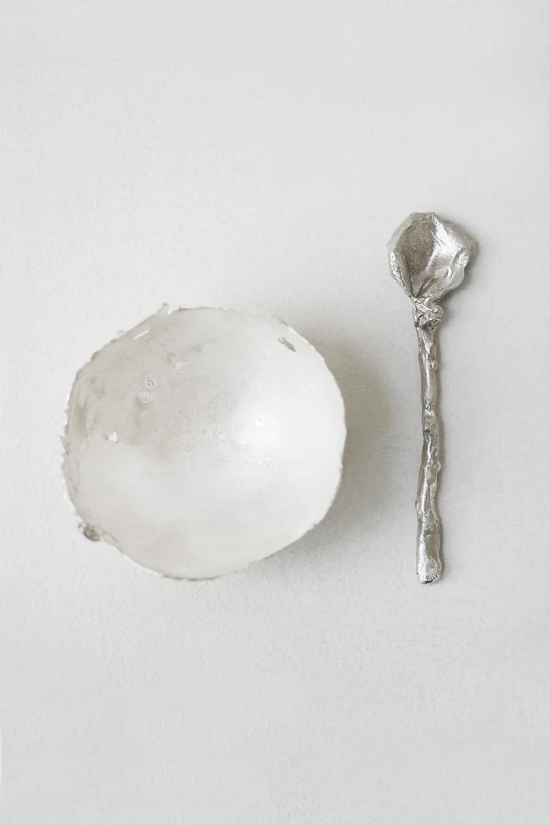 A textured white bowl sits beside the Completedworks L32 - Spoon in Silver, noted for its unique rough finish, on a light neutral background.