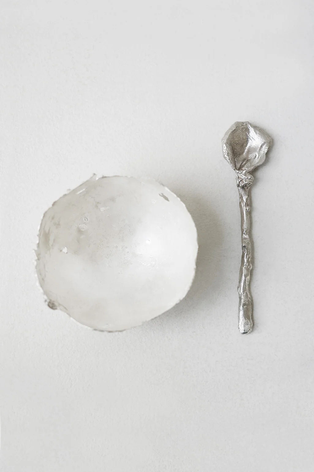 A textured white bowl sits beside the Completedworks L32 - Spoon in Silver, noted for its unique rough finish, on a light neutral background.