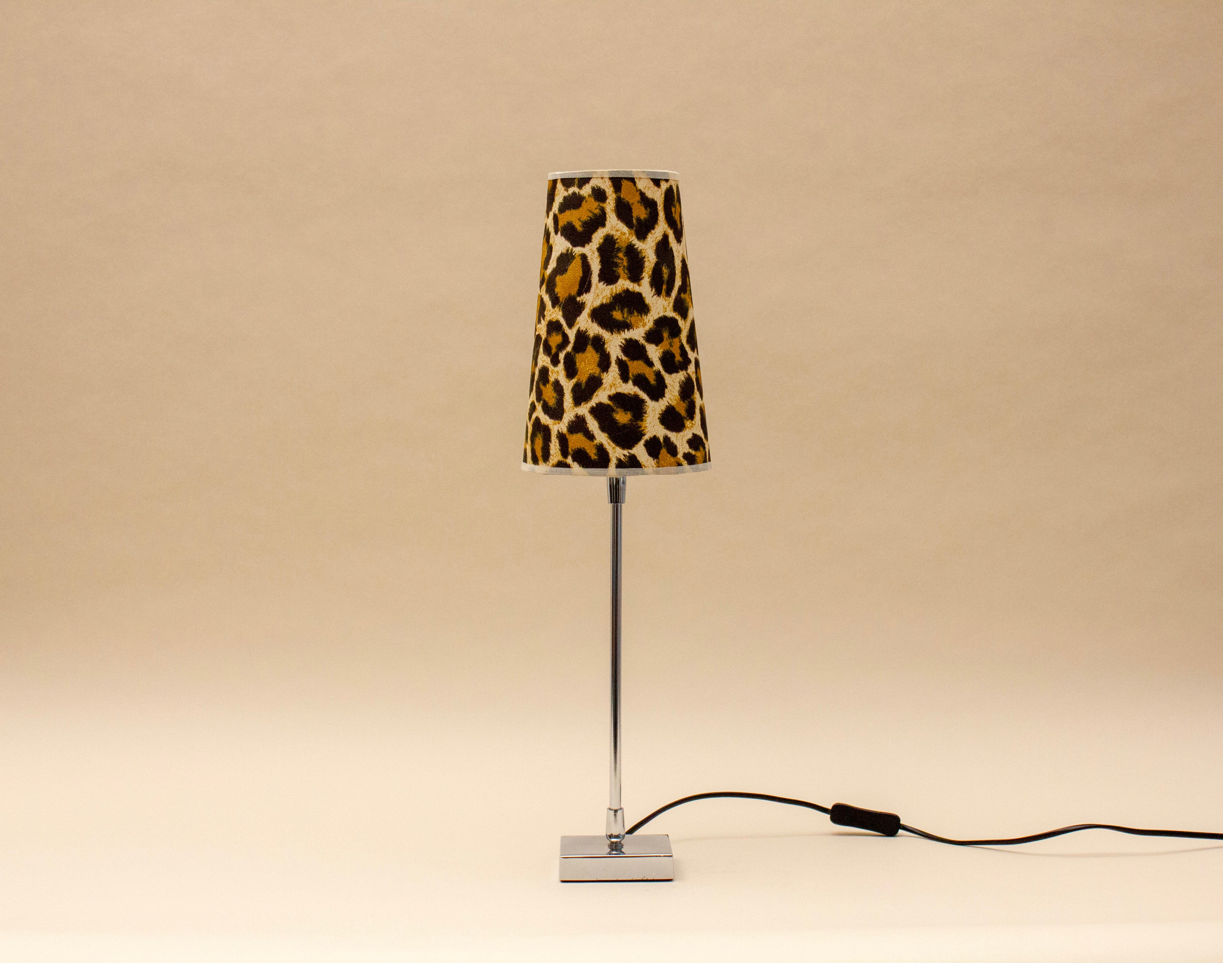 The SÒLER ZAFRA TABLE LAMP features a chrome base and a bold leopard print shade, resting on a neutral surface with its black power cord exposed. The plain beige background highlights its modern vintage design.