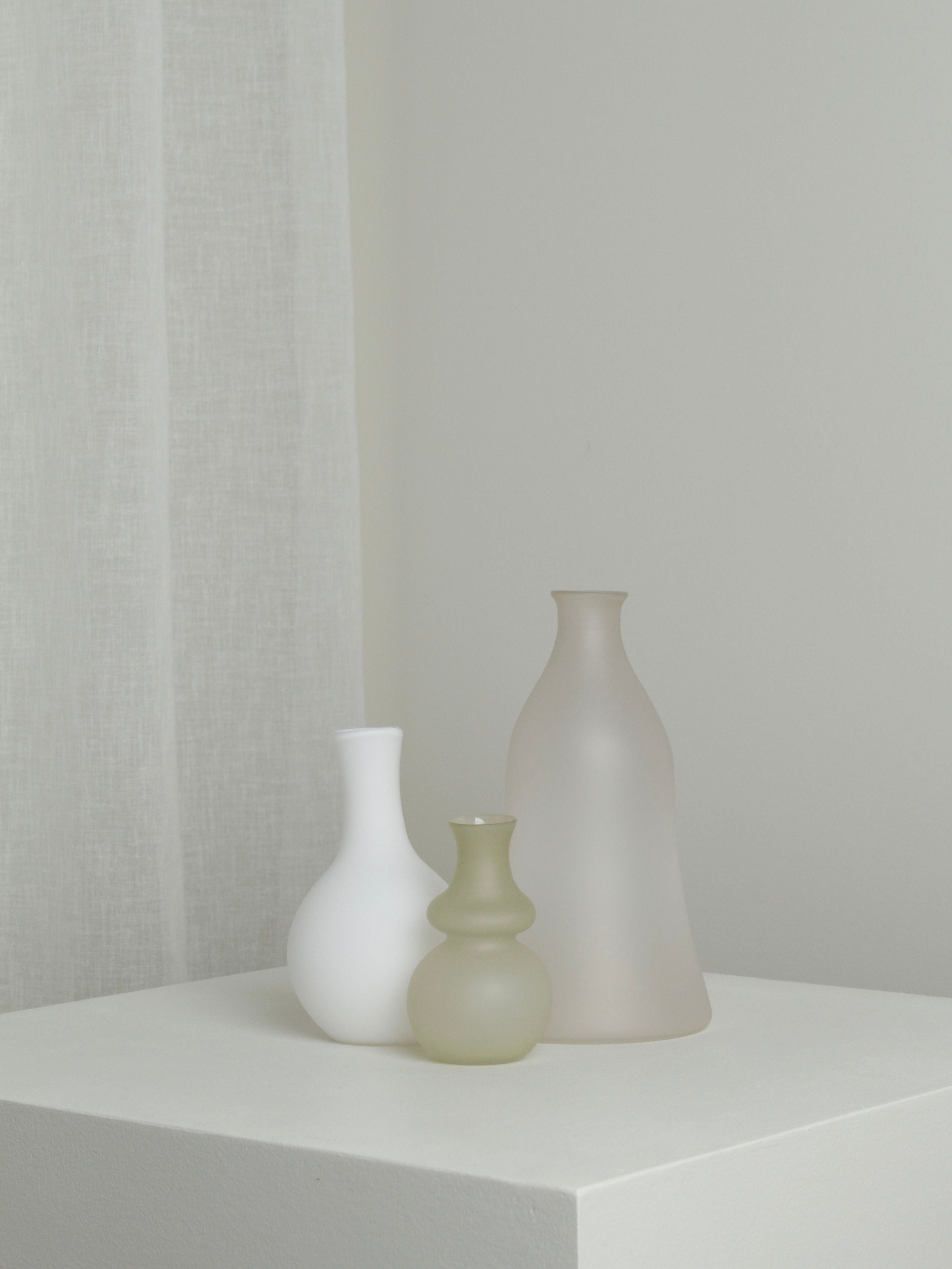 The Small Vase in Stone by Verre d'Onge stands on a white pedestal against a neutral wall and sheer curtain, creating a minimalist and serene display.