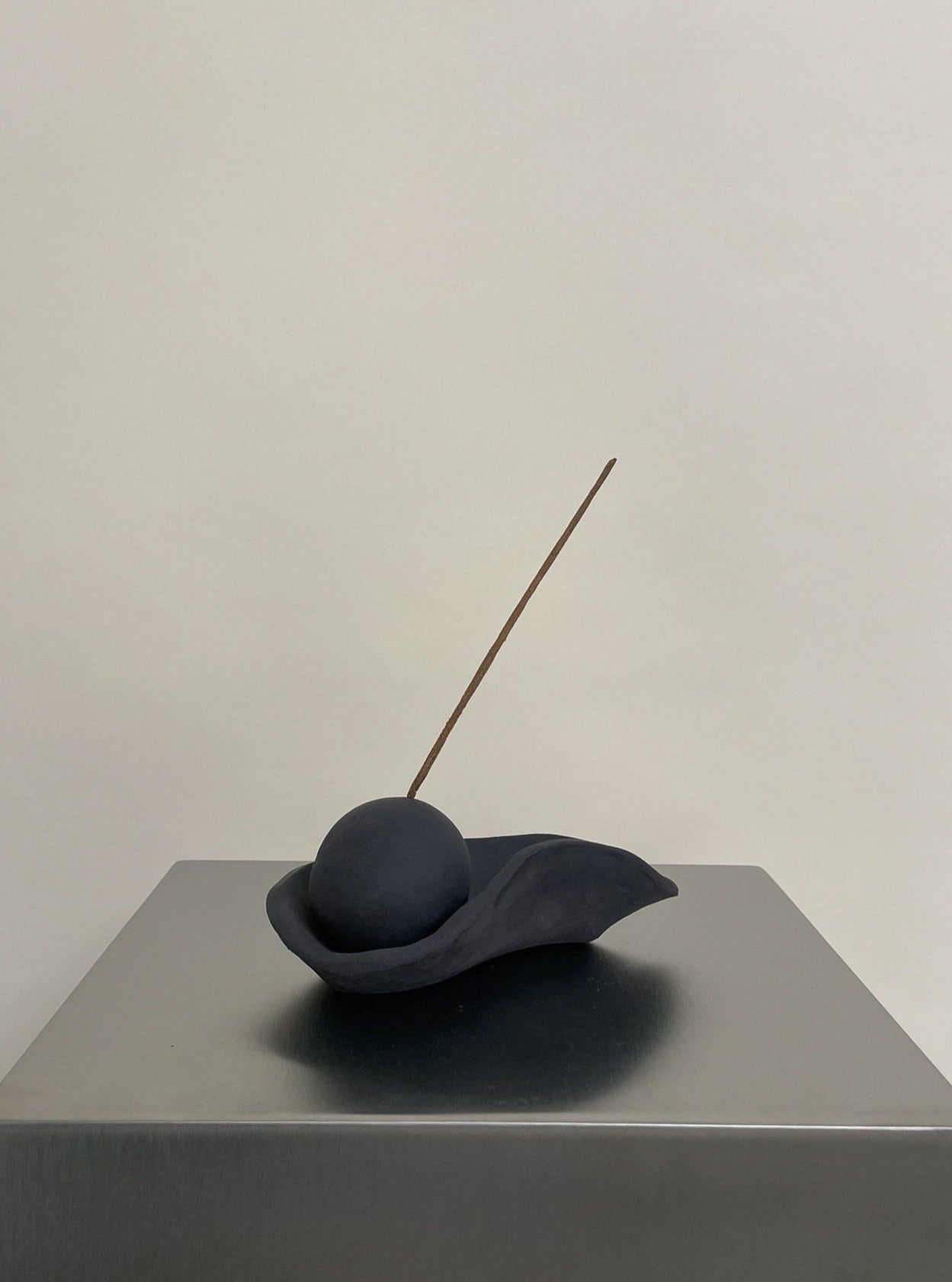 The Incense Holder black stoneware by 2222Studio features a dark, round sphere on an abstract leaf-shaped base with a thin incense stick—an elegant, minimalist accent for any home décor. Displayed on a smooth metallic surface against a plain background.