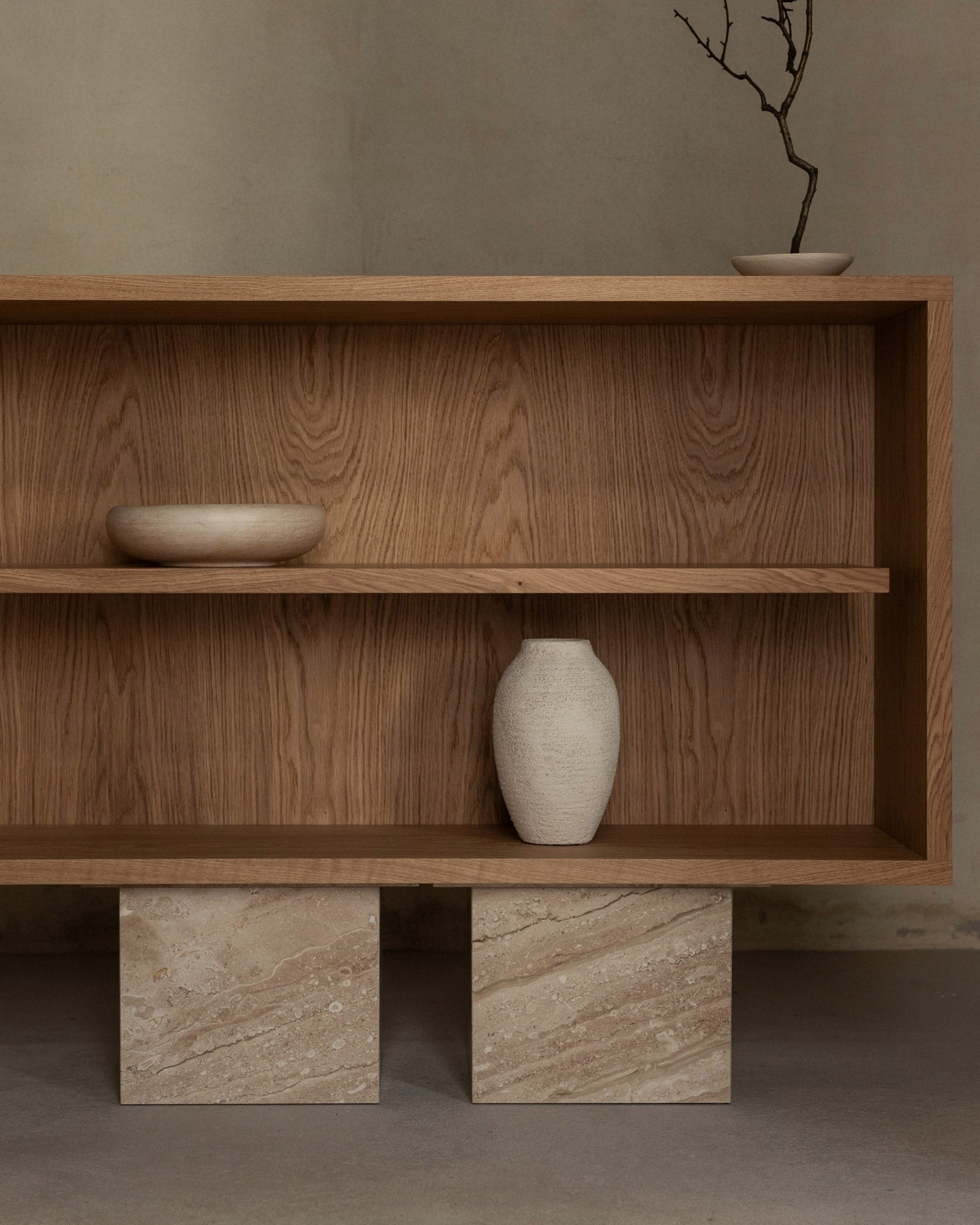 The BLOCK Sideboard by un'common, featuring minimalist wooden design, displays a round textured bowl, a white ceramic vase, and a shallow dish with a bare twig. It stands on two rectangular stone blocks against a neutral wall.