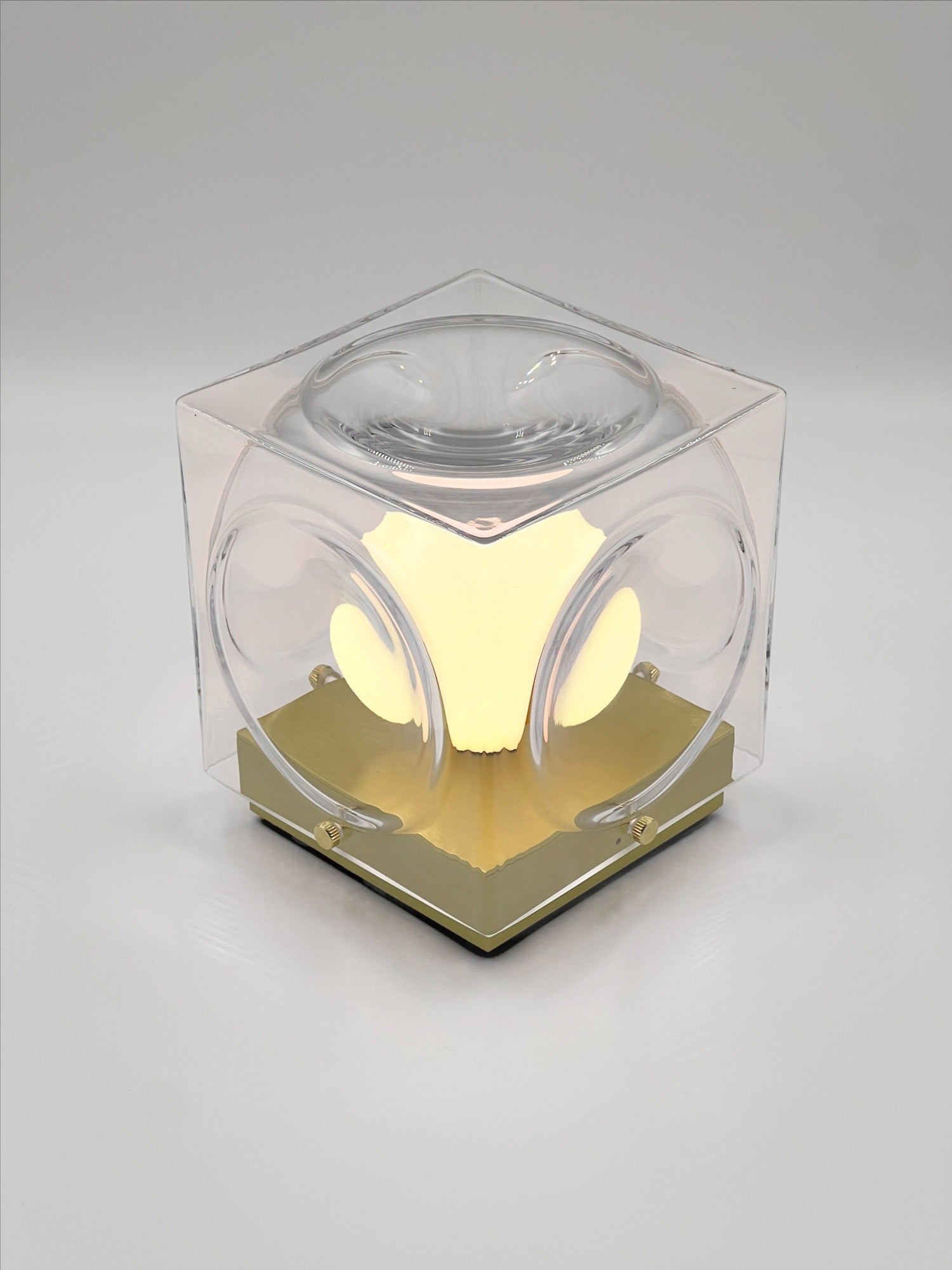 The Chant Portable Table Lamp by Lee Broom, a cube-shaped, weather-resistant lamp with a transparent shell and rounded side cutouts, emits a glowing yellow light as it sits on a flat surface against a plain background.