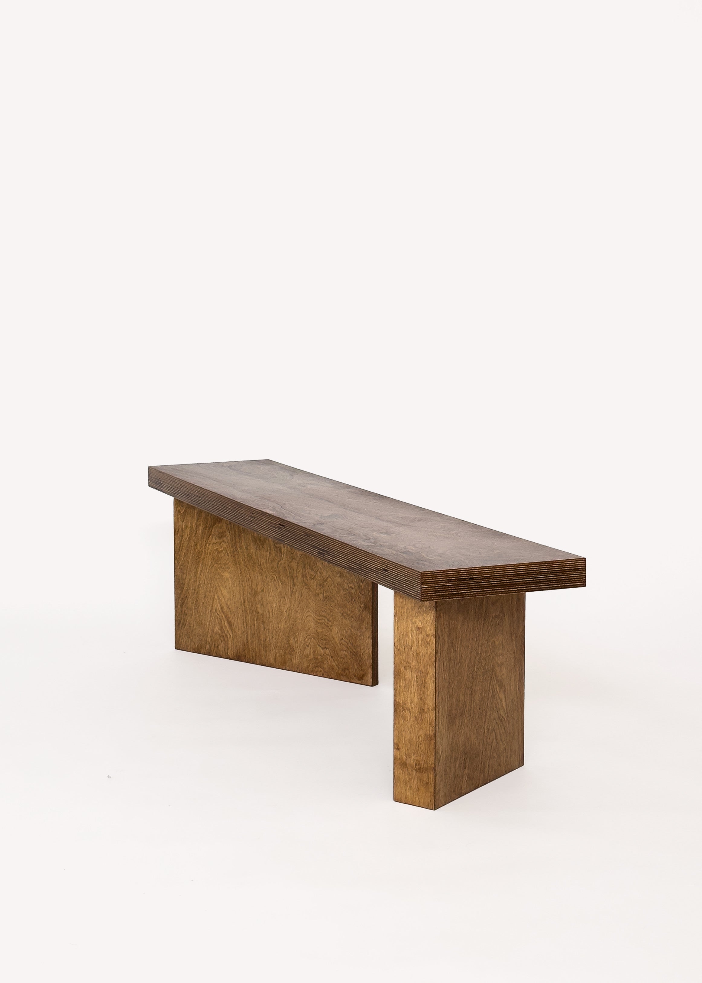 The Free plan Bench by GOONS features a customizable rectangular seat with two wide, solid legs in a natural brown finish. Its minimalist design, without backrest or armrests, is perfect for modern spaces. Set against a plain white background.