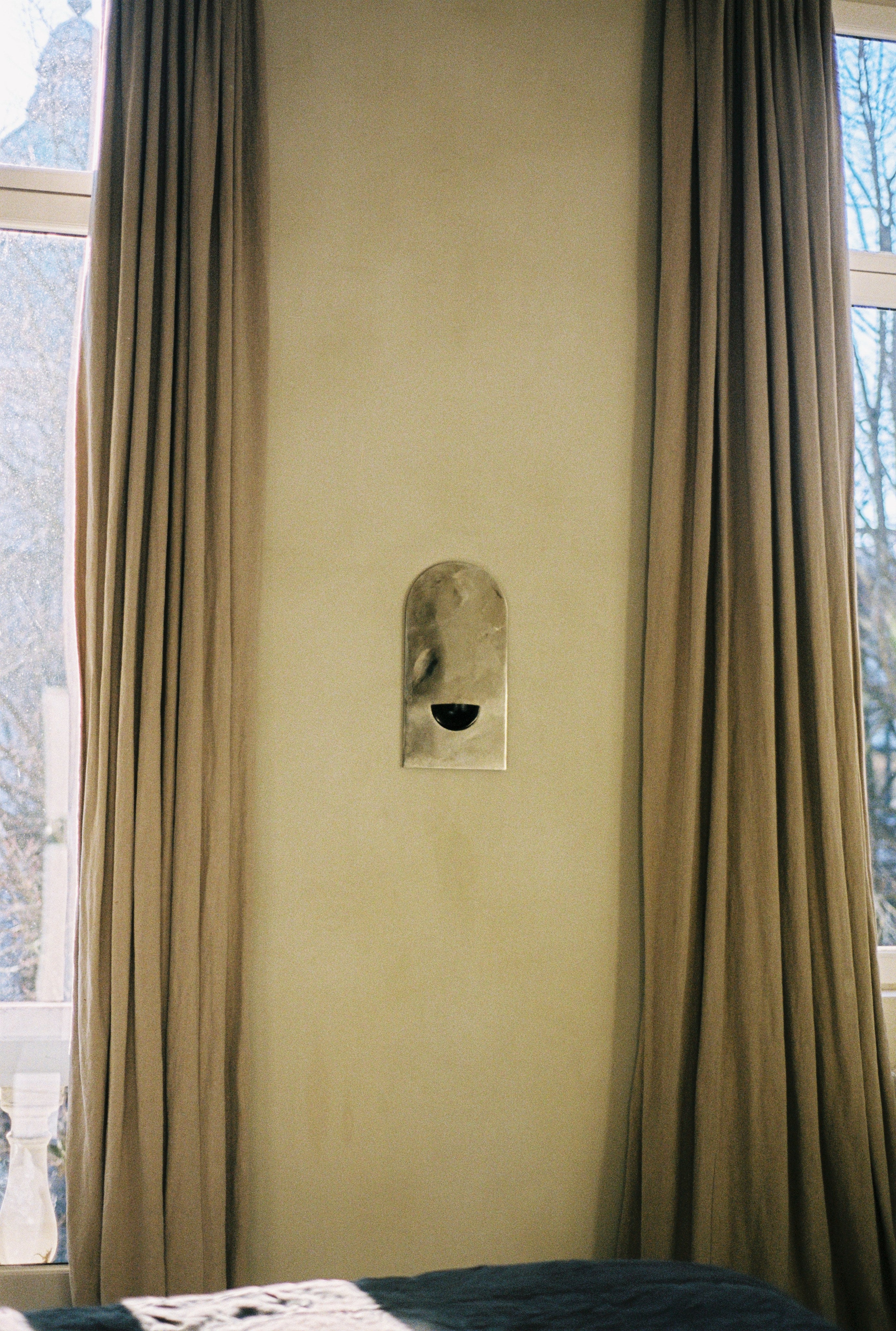 A beige wall with tall windows, framed by long, light brown curtains. Centered between them is The Mirror of Simple Souls, a contemporary steel mirror by COSEINCORSO—ideal for minimalist interior decor.