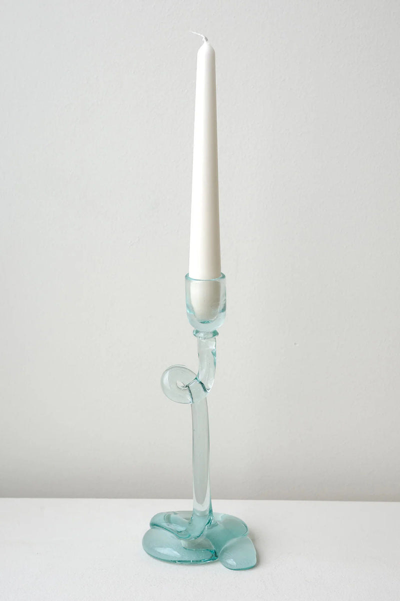 A white taper candle sits in the Thaw - Recycled Glass Candleholder in Clear by Completedworks, featuring an abstract wavy design, placed on a white surface with a plain light gray background.