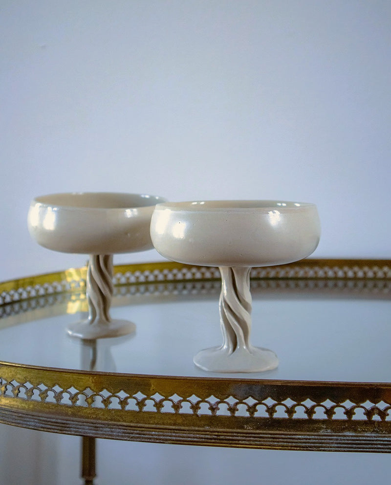 Two cream-colored Ceramic Champagne Coupe Contemporary goblets by Anna Karountzou, featuring twisted stems, rest on a round, gold-edged glass table against a plain light background.