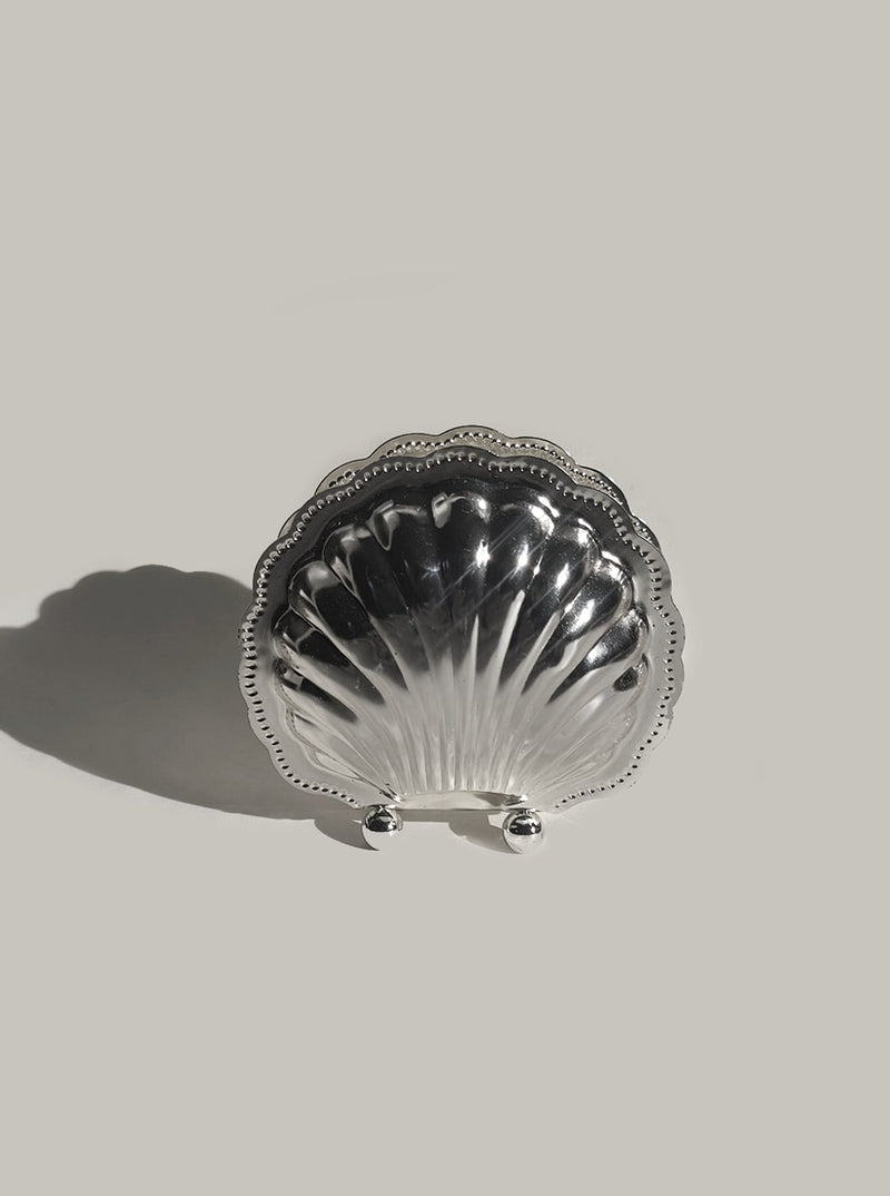 The Les Objoies Napkin Shell Holder is a silver-plated, seashell-shaped napkin holder with scalloped edges and two round feet, displayed upright against a plain light gray background.