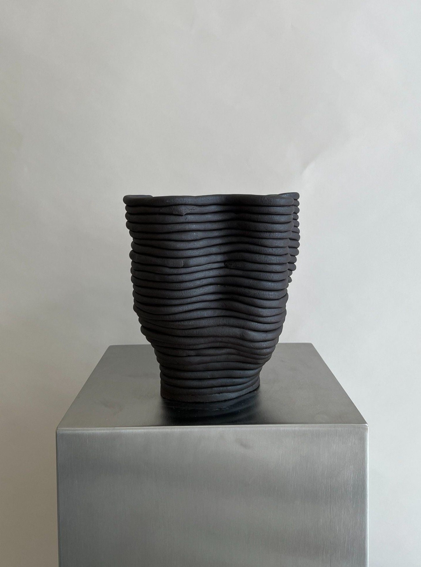 The Wobbly Vase Black by 2222Studio, featuring a dark wavy form and ribbed, layered texture, stands on a reflective metal pedestal against a light gray background for a refined decorative touch.