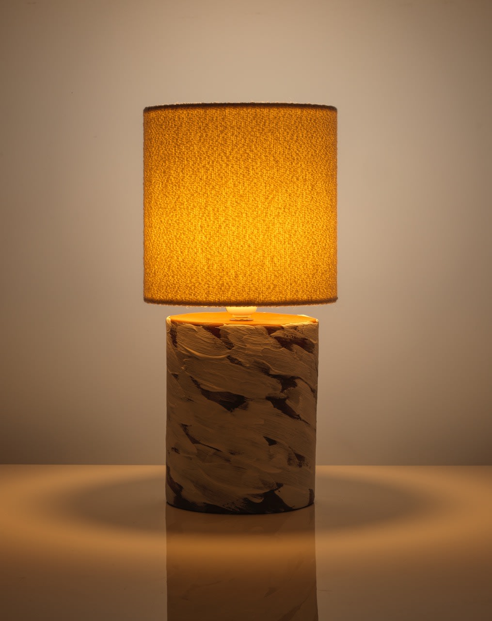 The White Wind Table Lamp by Katia Langeard features a lit, cylindrical textured yellow shade and marbled ceramic base, resting on a reflective surface and casting a warm glow against a plain backdrop.