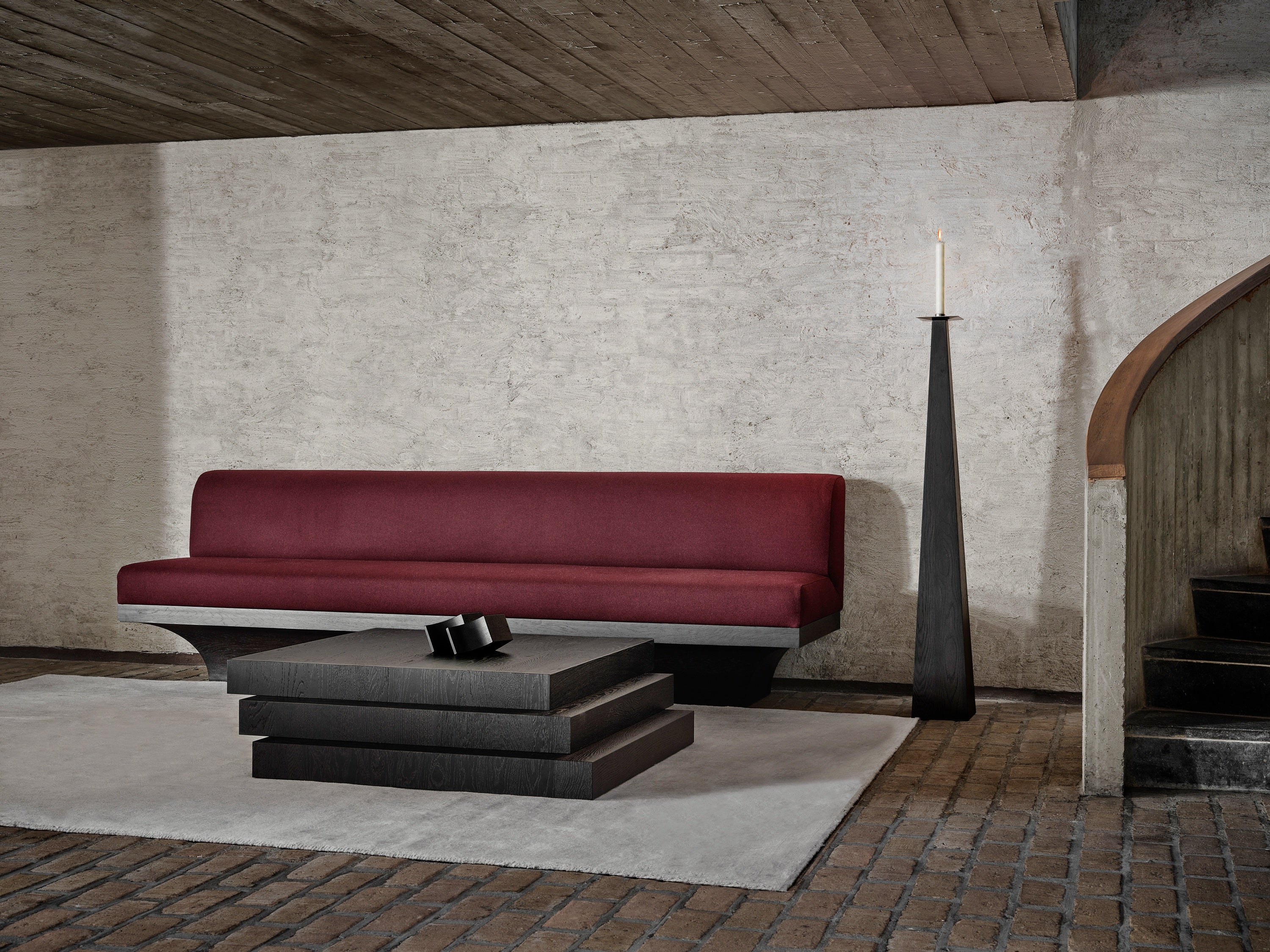 A modern living room features the Mokko Acer Sofa in deep red, a stack-style black coffee table, a tall black candle holder, and a white rug on brick floors. Textured walls and a wooden staircase complete this inviting space.