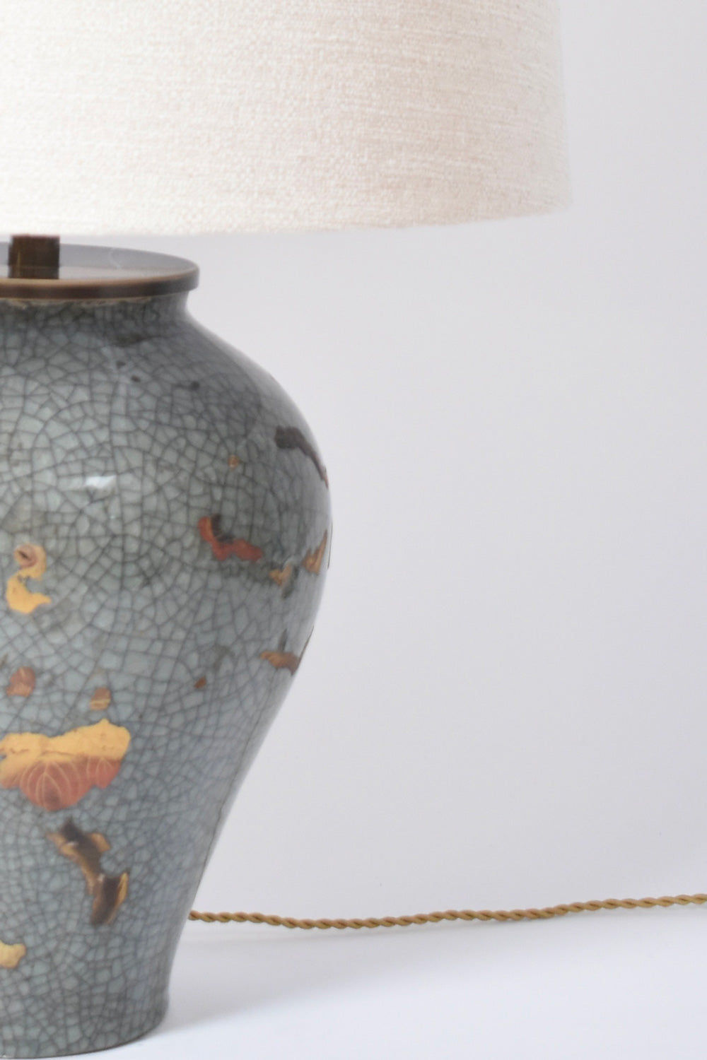 The Chinese Celadon and Gold Lamp, XIXth c. by Barracuda Interiors features a crackled gray and yellow design, a textured white lampshade, and a twisted gold cord, displayed on a white surface.