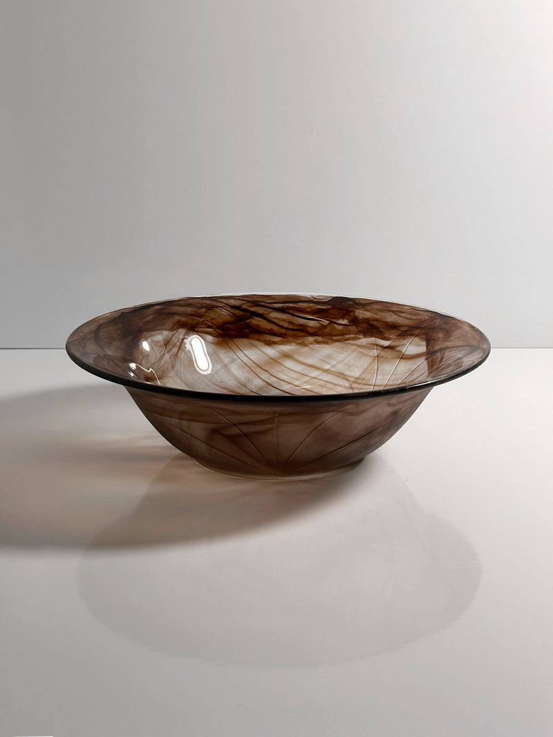 The Dodo Vintage German Art Deco Cloud Glass Bowl has a shallow, round shape with a wide rim and swirled brown and smoky patterns, evoking vintage style. It's displayed on a smooth white surface against a plain light background.