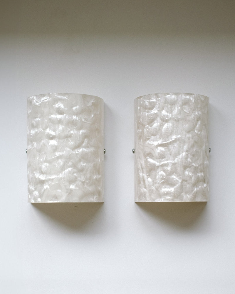 A Set of 2 Mother-of-pearl Wall Lamps by septembre studios, featuring a wavy, hammered texture and semi-cylindrical white design reminiscent of 1980s style, are mounted side by side on a light-colored wall.