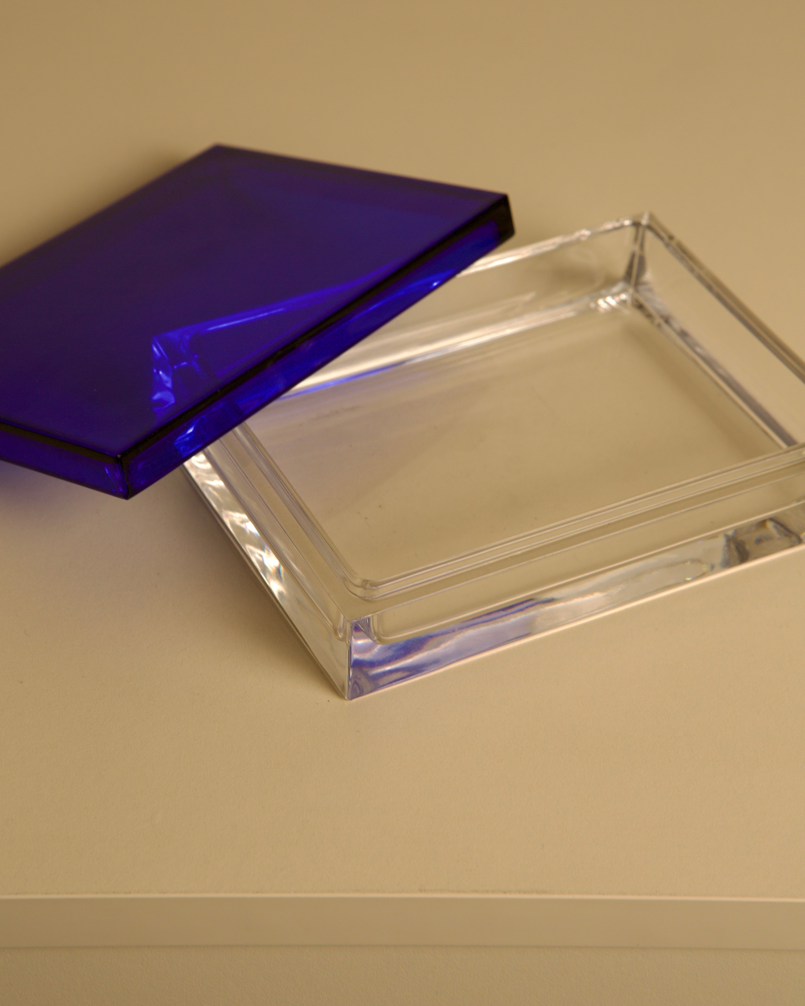 The Italian Box in Transparent and Blue Crystal by Duccio Di Segna 80's from Treaptyque features a rectangular form with a blue-tinted lid, partially open on a beige surface, reflecting iconic 1980s Italian design.