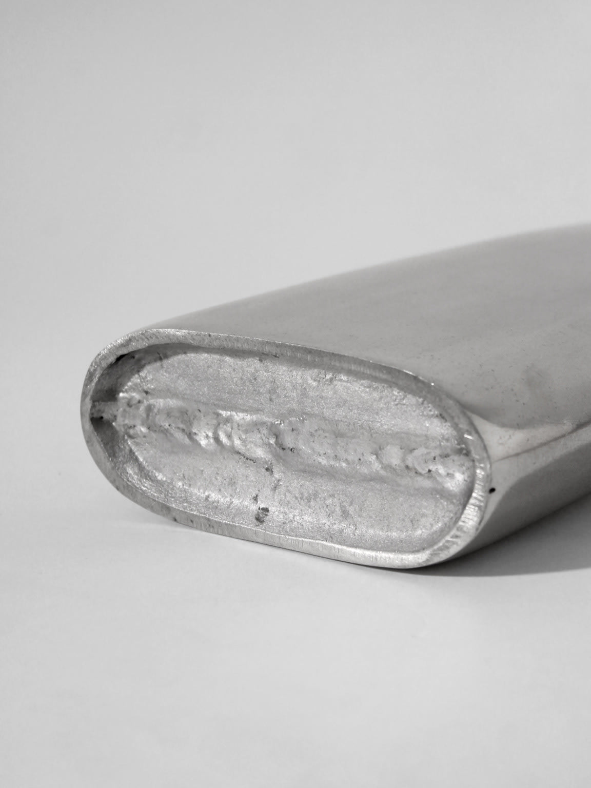 A close-up of the Polished Aluminum Flat Tall Vase by Vintage Stories, lying on its side against a plain white background. The vase’s shiny, oval form and textured metallic surface are clearly visible.