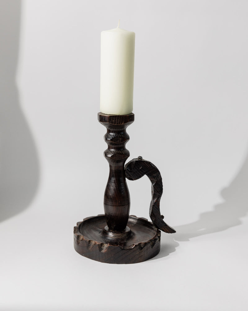 A Bottega Jacobs 1960s wooden candleholder in handcarved dark wood holds a white candle upright, featuring a decorative curved handle, set against a plain white background.